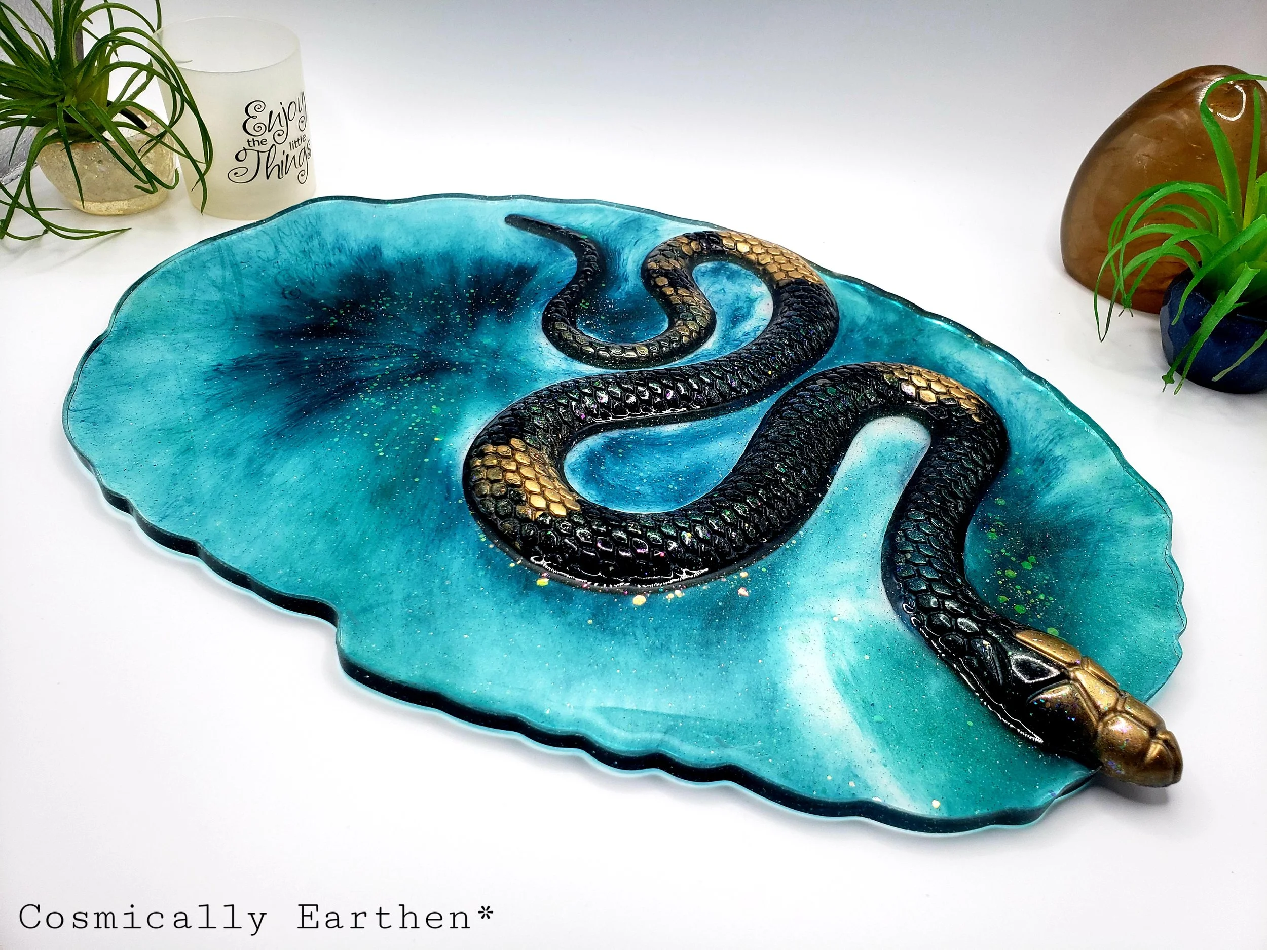 Handcrafted resin geode tray in vibrant blue with a black and gold snake design. Crystals rest on the tray, which is used for displaying sacred items. Surrounded by plants and mystical decor.