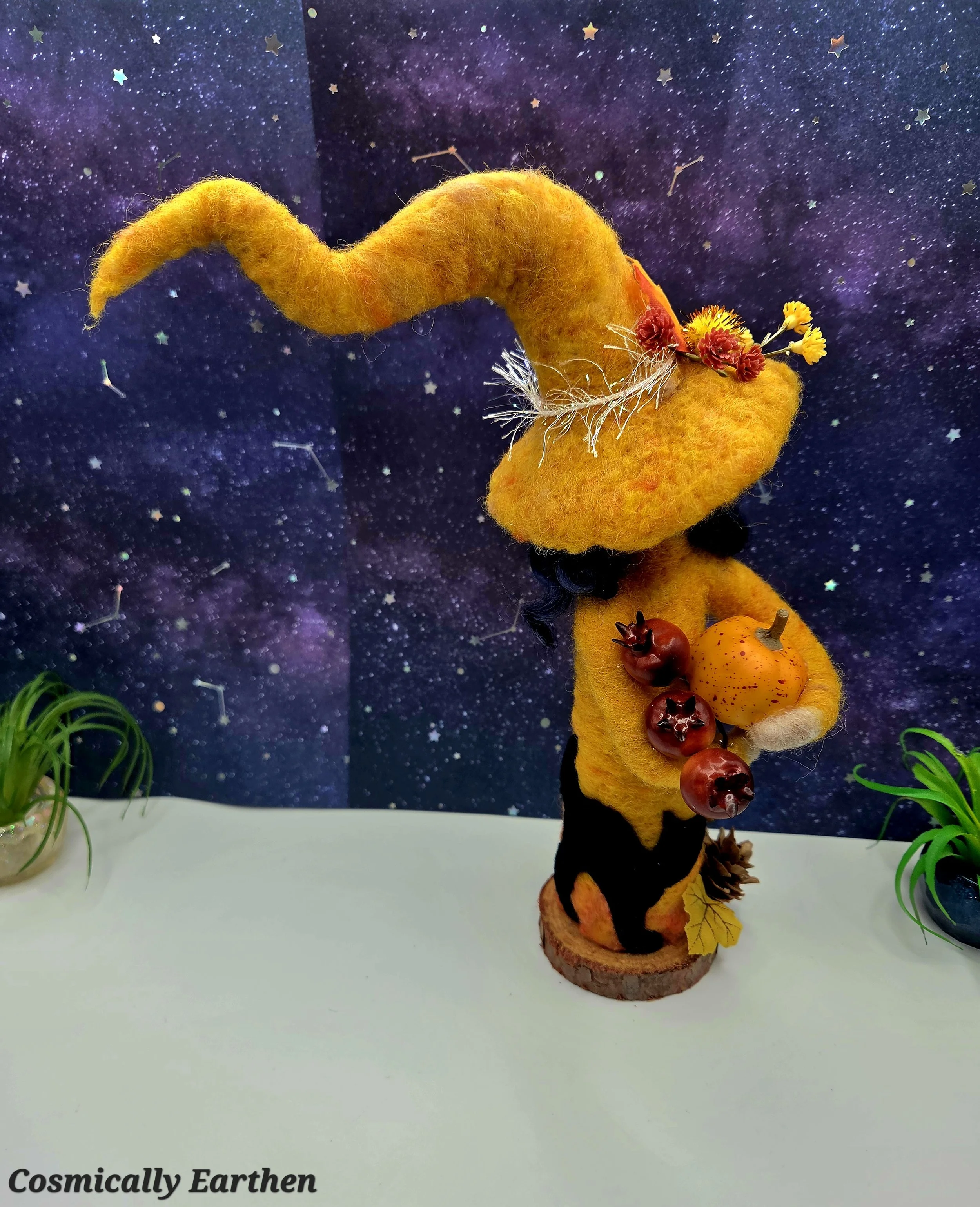 Handcrafted needle felted fall witch doll in warm orange tones with a curved hat, holding a pumpkin and pomegranates, detailed with wool textures and mounted on a natural wood base as a collectible fiber art sculpture