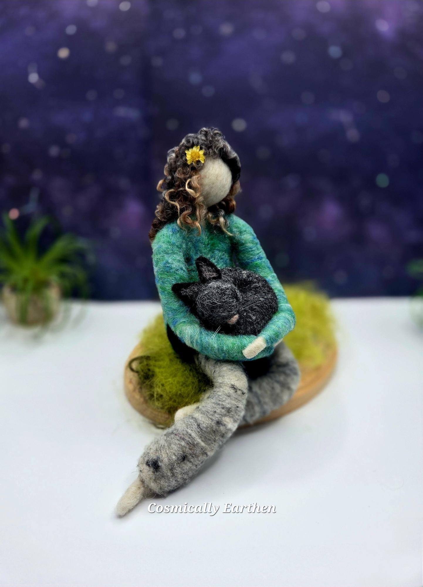 Needle felted doll with curled hair holding a sleeping black cat, seated on a soft green grass cloud base with a dreamy purple background