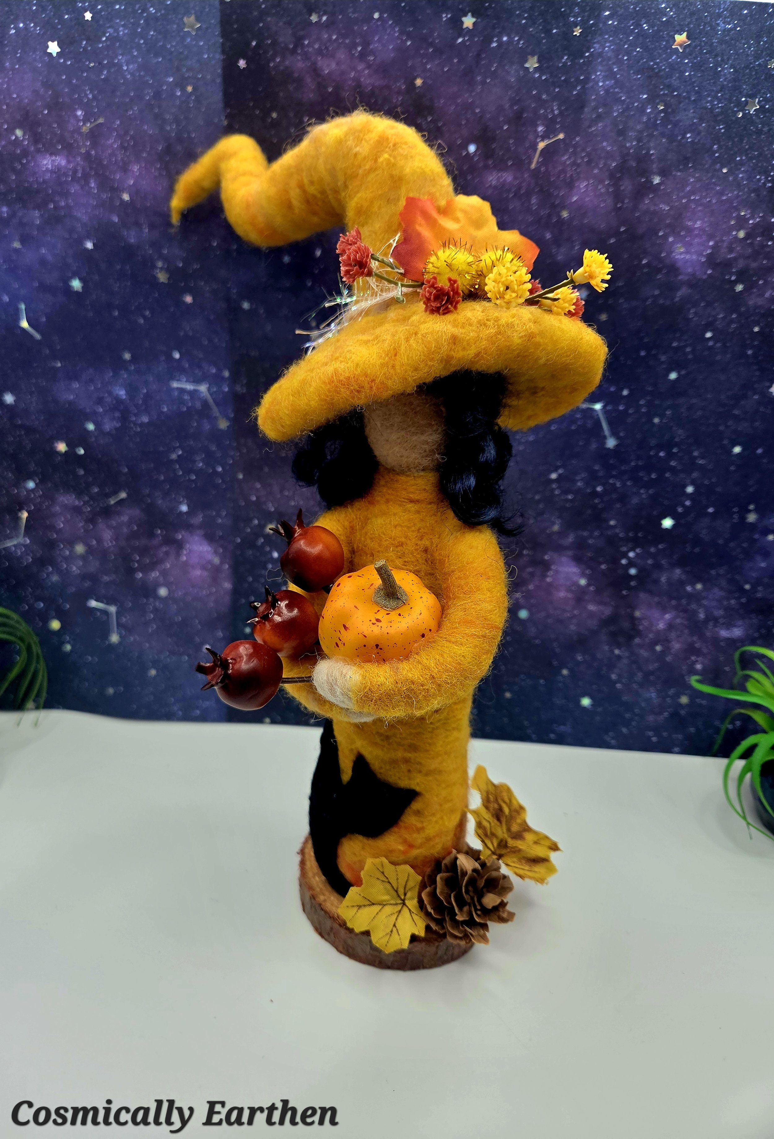 Side view of handmade needle felted autumn witch doll Marigolde with layered wool clothing, curved hat, and harvest elements, displayed on a wood base as a collectible fiber art sculpture