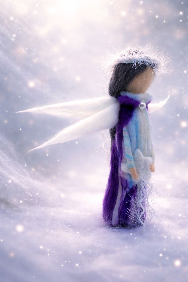 Handcrafted needle-felted angel doll shown in profile, with soft white wings, a shimmering halo, and flowing blue and purple wool garments, standing in a snowy, luminous setting with gentle sparkles.