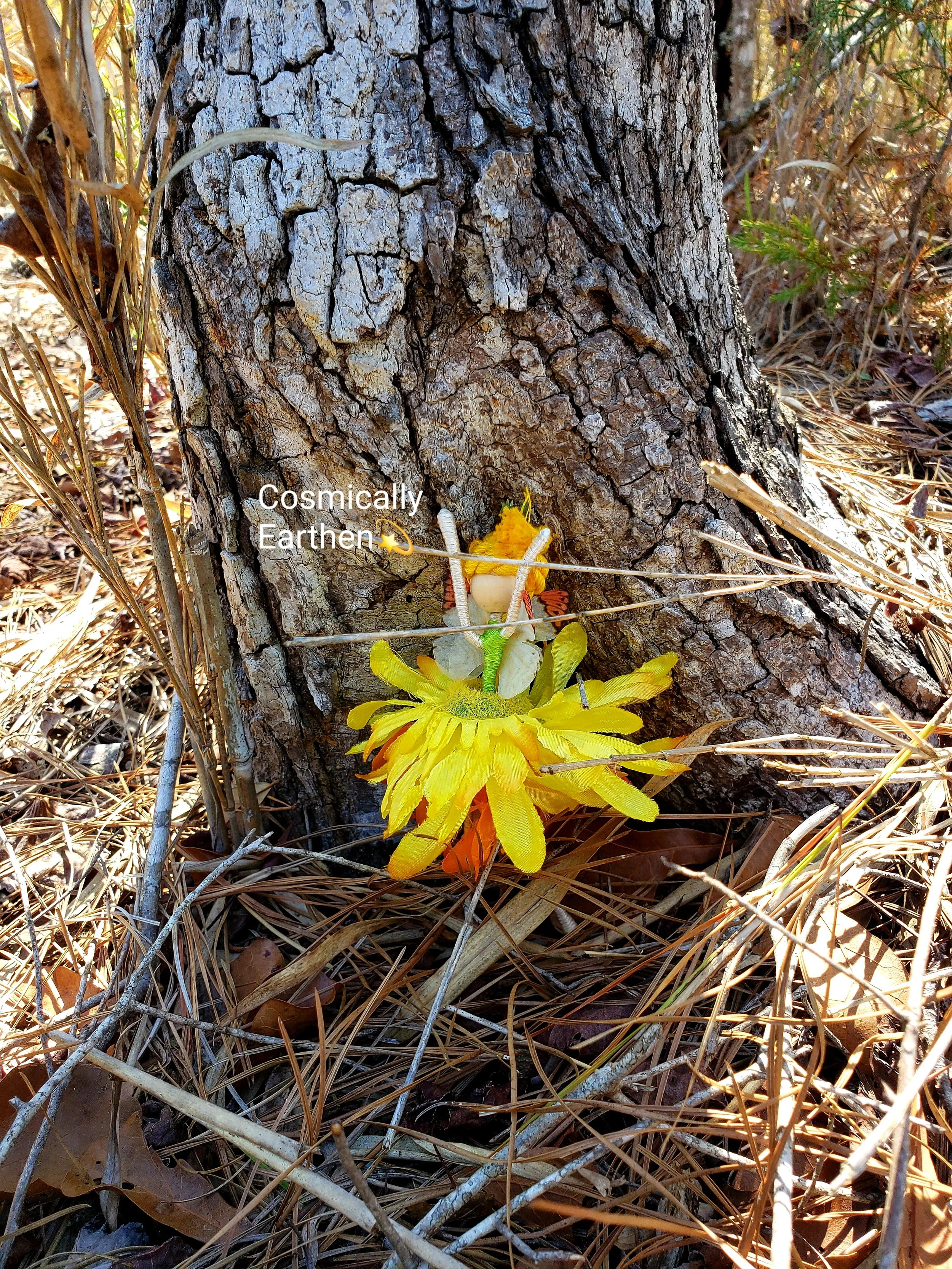 A small handcrafted fairy with yellow flower petals resting at the base of a tree trunk among pine needles and fallen leaves in a forest setting.