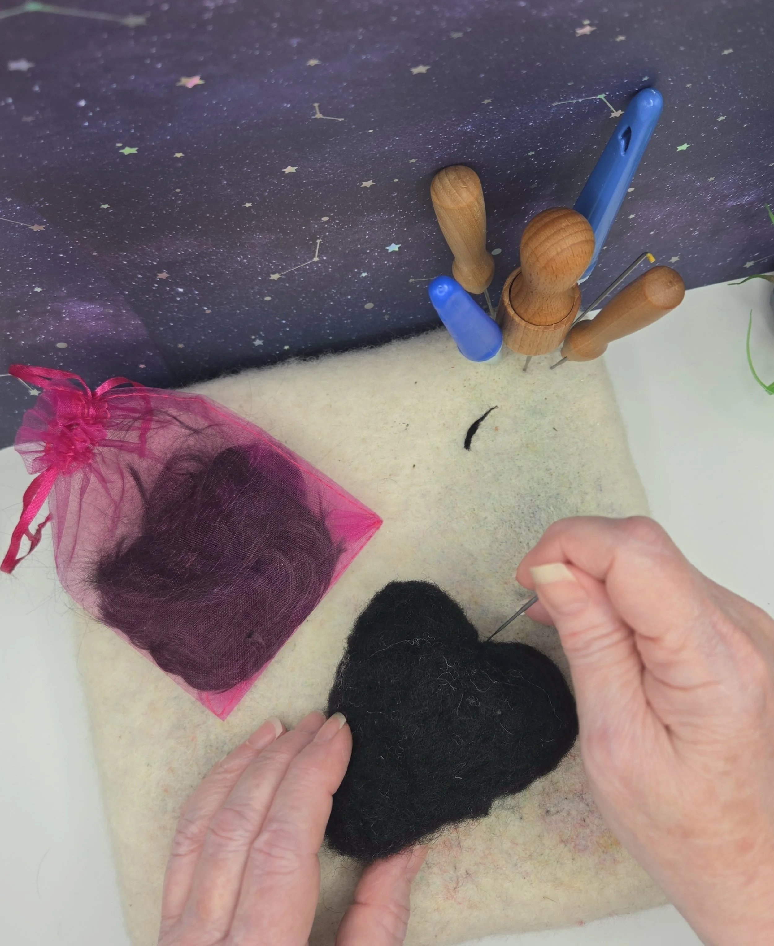 A person sewing a black felt heart with a needle. Two felt hearts, one black and one pink, are on a white surface. A pink organza bag with black felt inside is nearby. Embroidery tools are in a wooden stand, and a starry night sky backdrop is behind.
