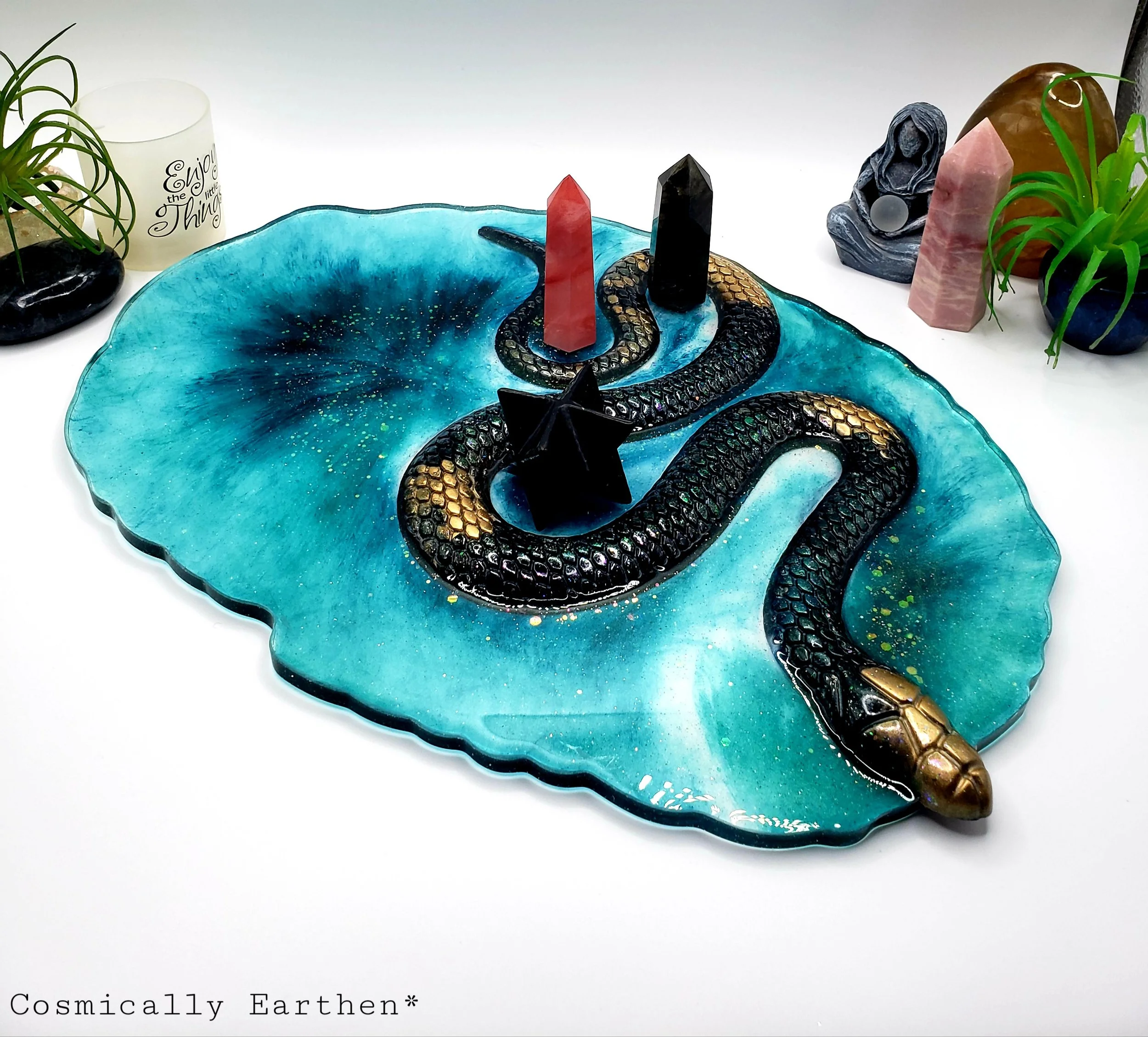 Handcrafted resin geode tray in vibrant blue with a black and gold snake design. Crystals rest on the tray, which is used for displaying sacred items. Surrounded by plants and mystical decor.