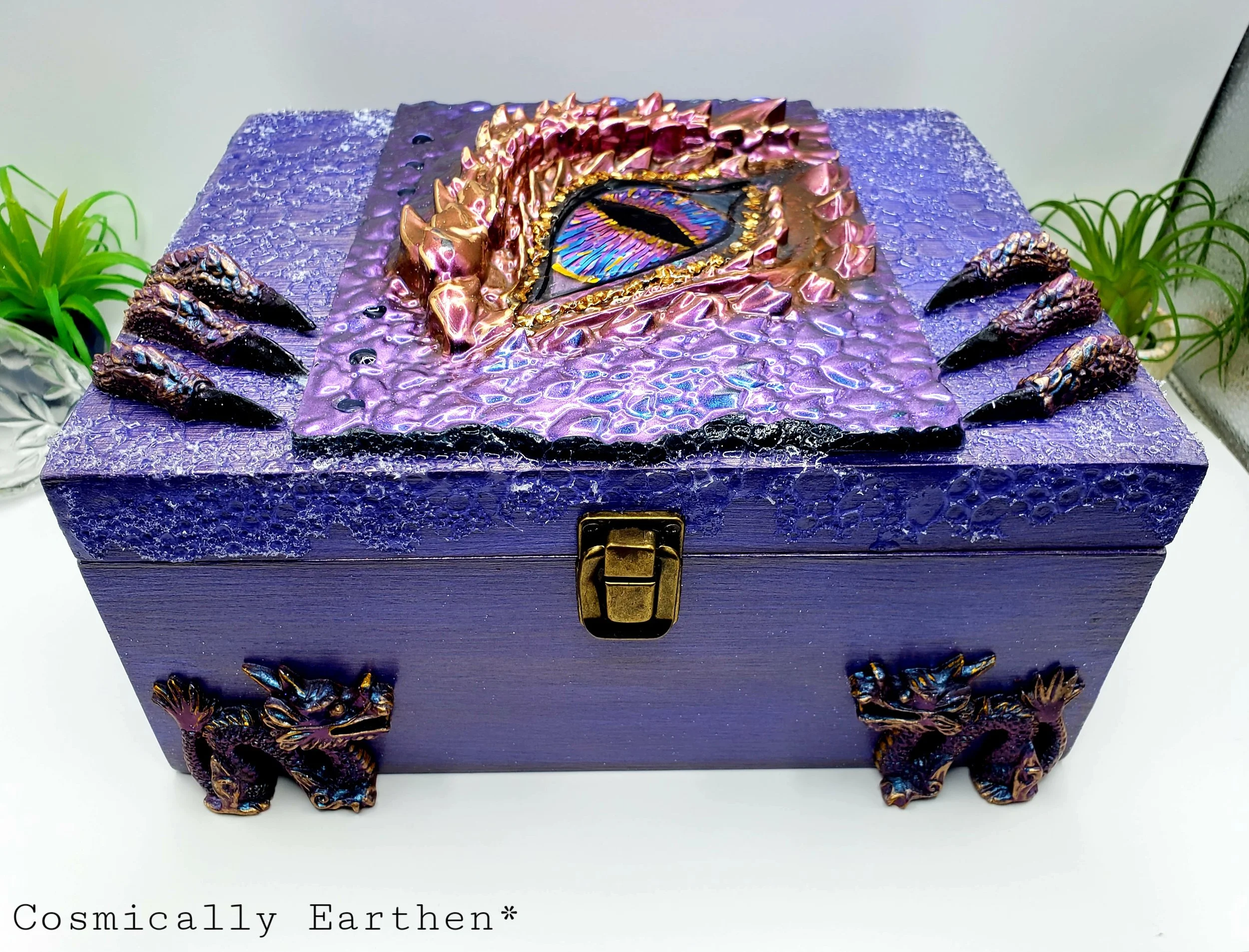 Decorative purple jewelry box with a stunning, colorful eye design on the lid, surrounded by metallic, flower-like embellishments, placed against a white background with green plants on either side.