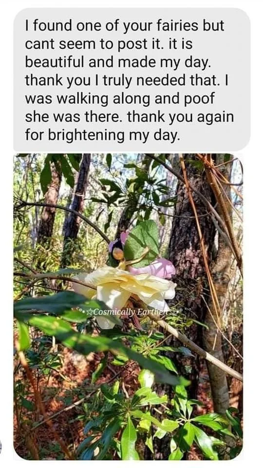 A white flower and a fairy figurine are nestled among trees and green foliage in a forest setting.