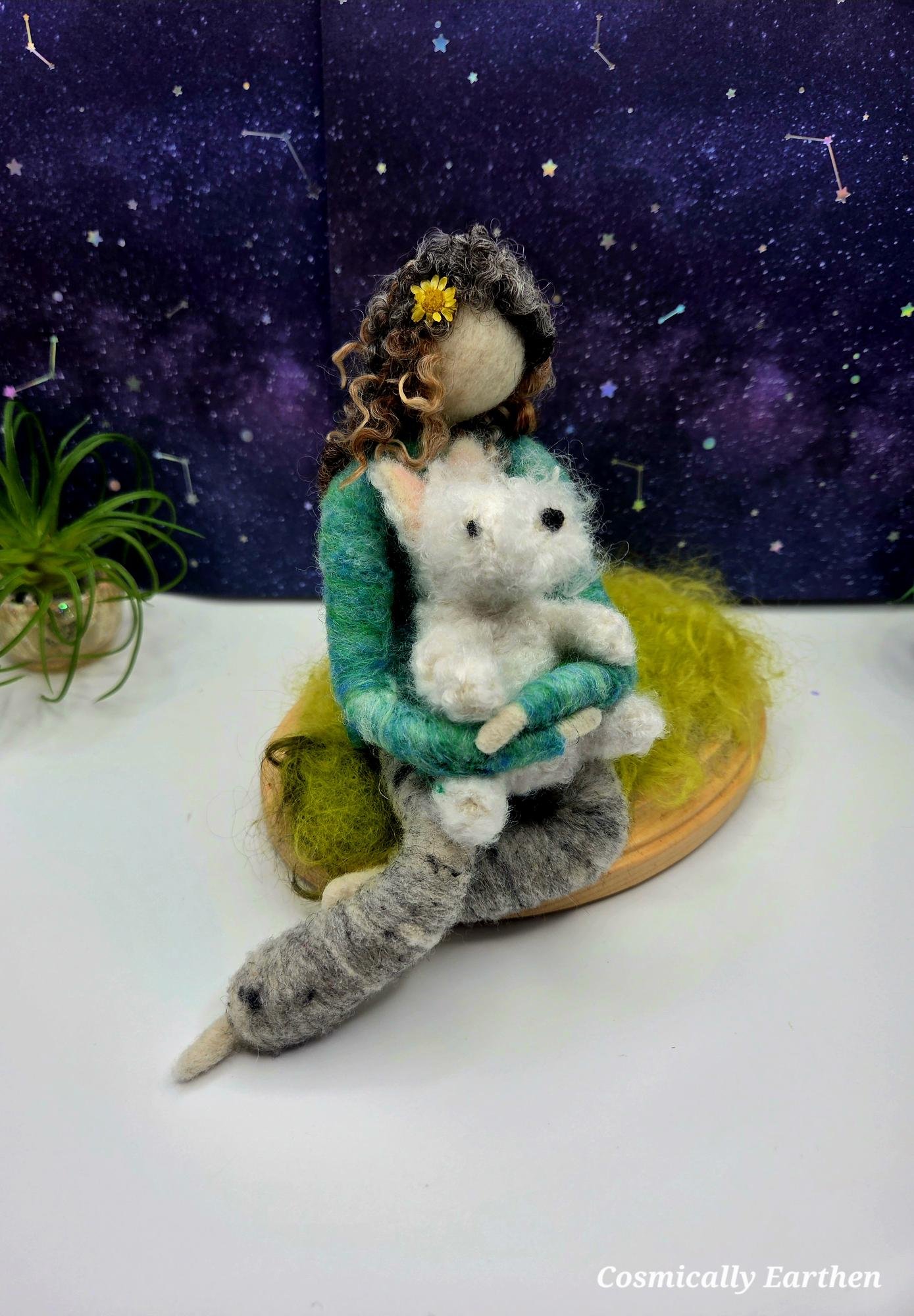 A handmade felt doll with curly hair holding a felt cat, sitting on a wooden base against a starry galaxy background.