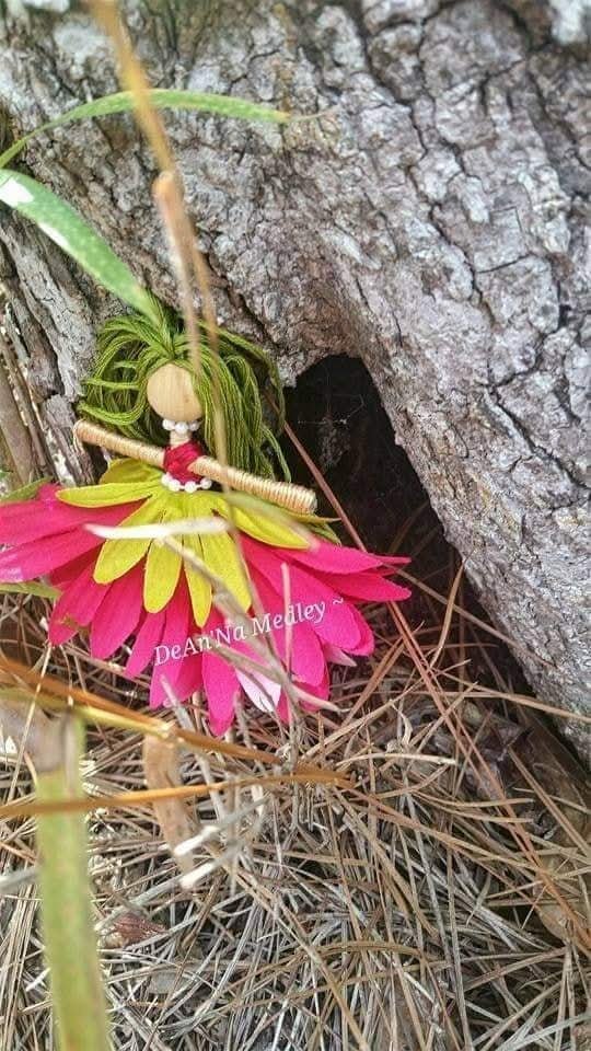 A handmade fairy doll with green hair, red and white outfit, sitting on pink and yellow flower petals, next to a tree trunk with a hole in the bark and dry grass on the ground.