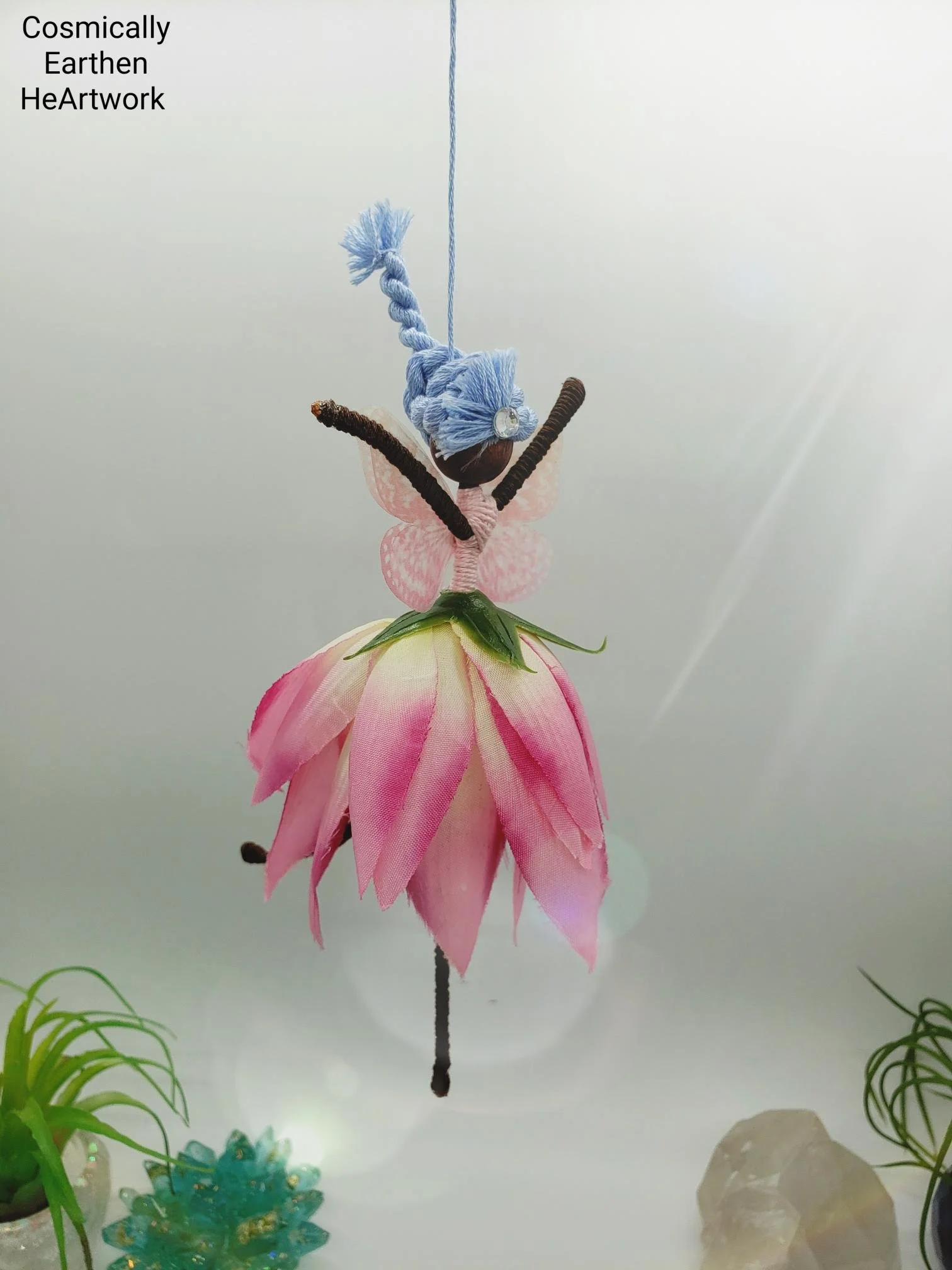 A craft of a butterfly made with fabric and other materials, hanging from a string, resembling a flower and plant scene on a white background.