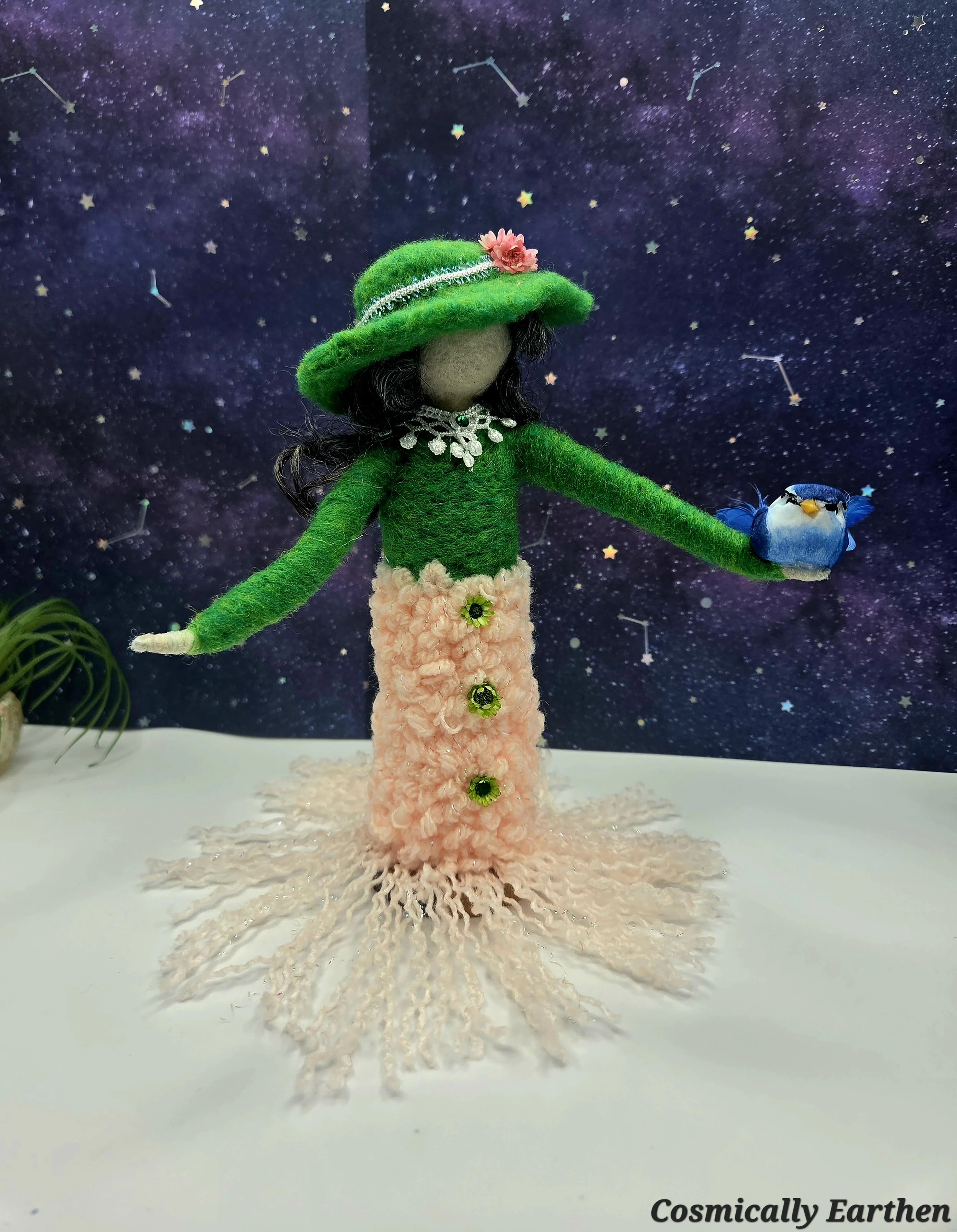 Handmade needle felted green doll Liora holding a small bird, with layered wool textures and earthy tones, standing on a natural wood base as a one-of-a-kind fiber art figure