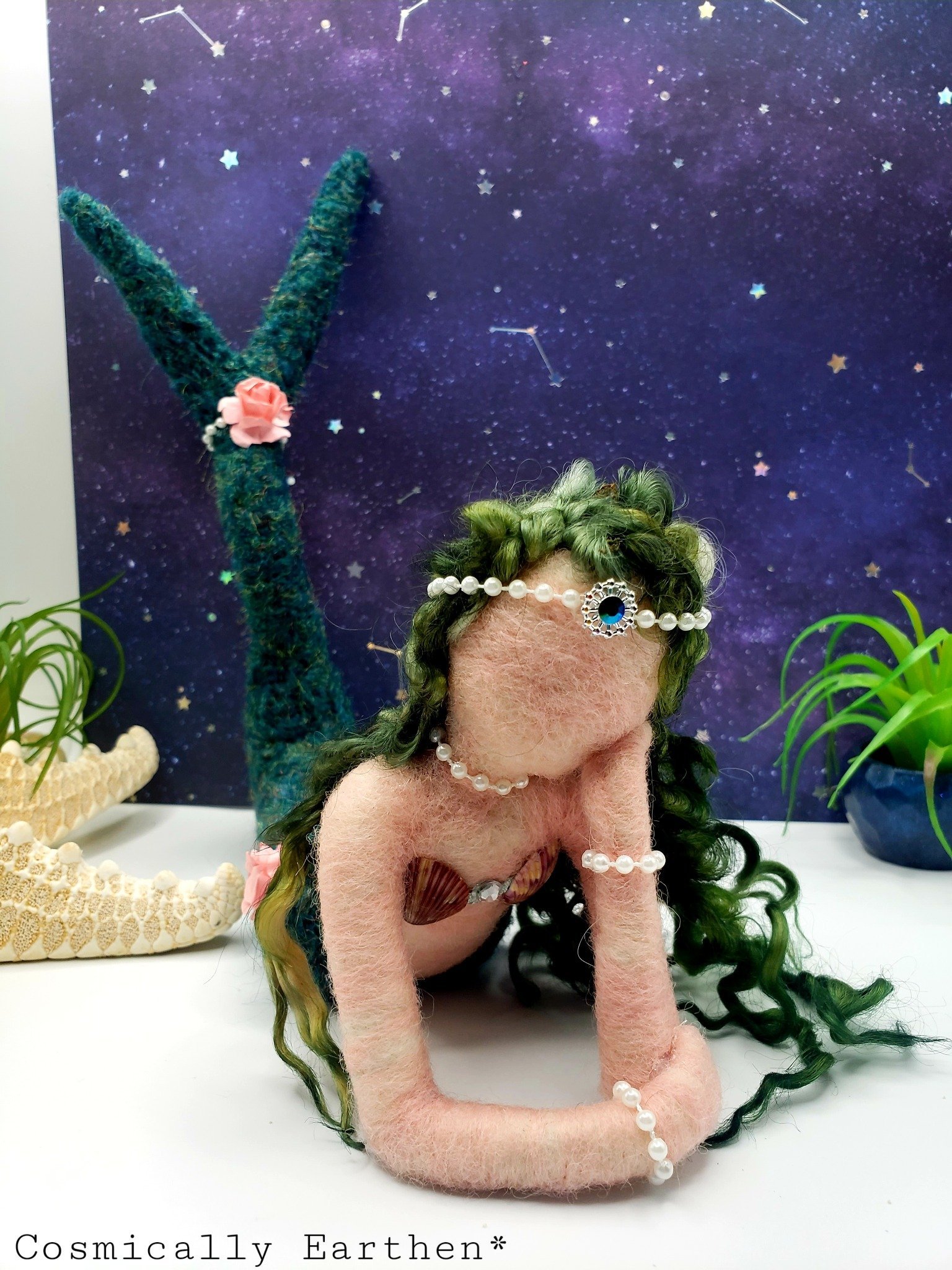 Felted mermaid sculpture with green curly hair, blue gemstone, and pearl jewelry, posed on a white surface against a space-themed purple background with star and constellation decorations.