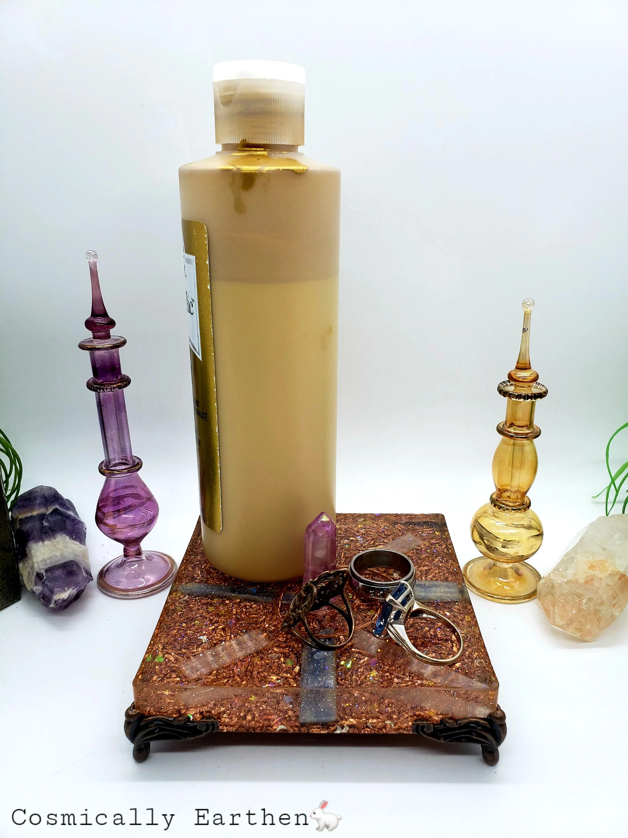 Handcrafted ethereal orgonite activator with pink AB quartz point, copper shavings, kyanite, and selenite, resting on four ornate feet with surrounding crystals and glass perfume bottles. Image 2: