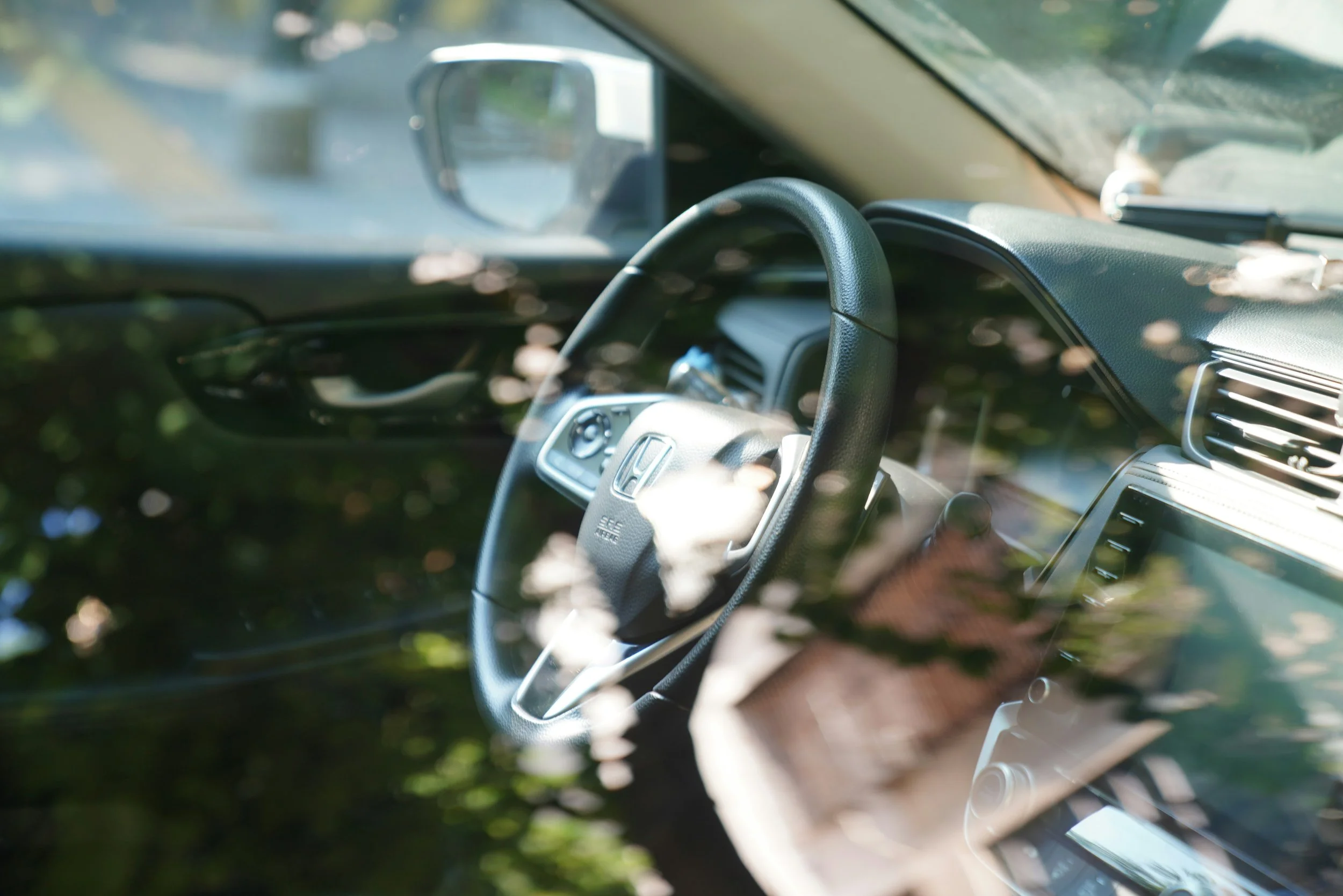 View of a car interior seen through the windshield, showing a steering wheel with a Honda logo, dashboard, and side mirror, with reflections of trees outside.