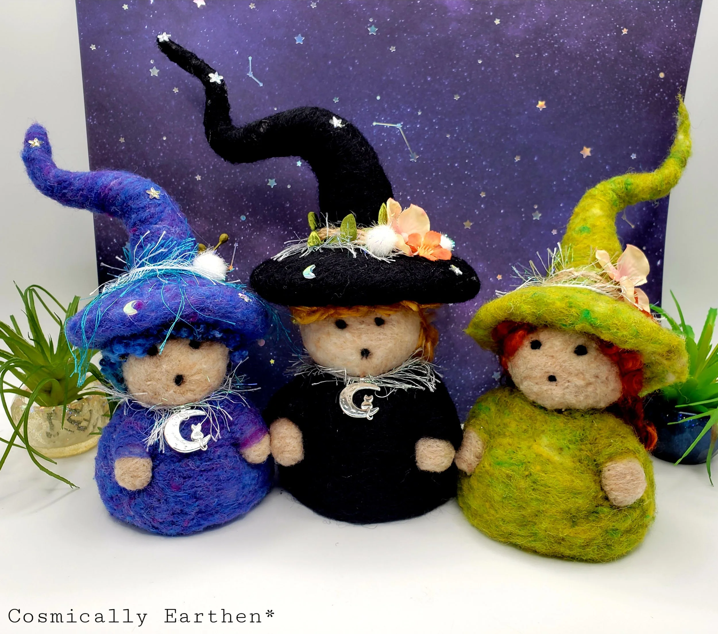 Needle-felted Groundling trio with blue, black, and green hats, adorned with flowers, crystals, and charms, set against a starry background with small green plants.