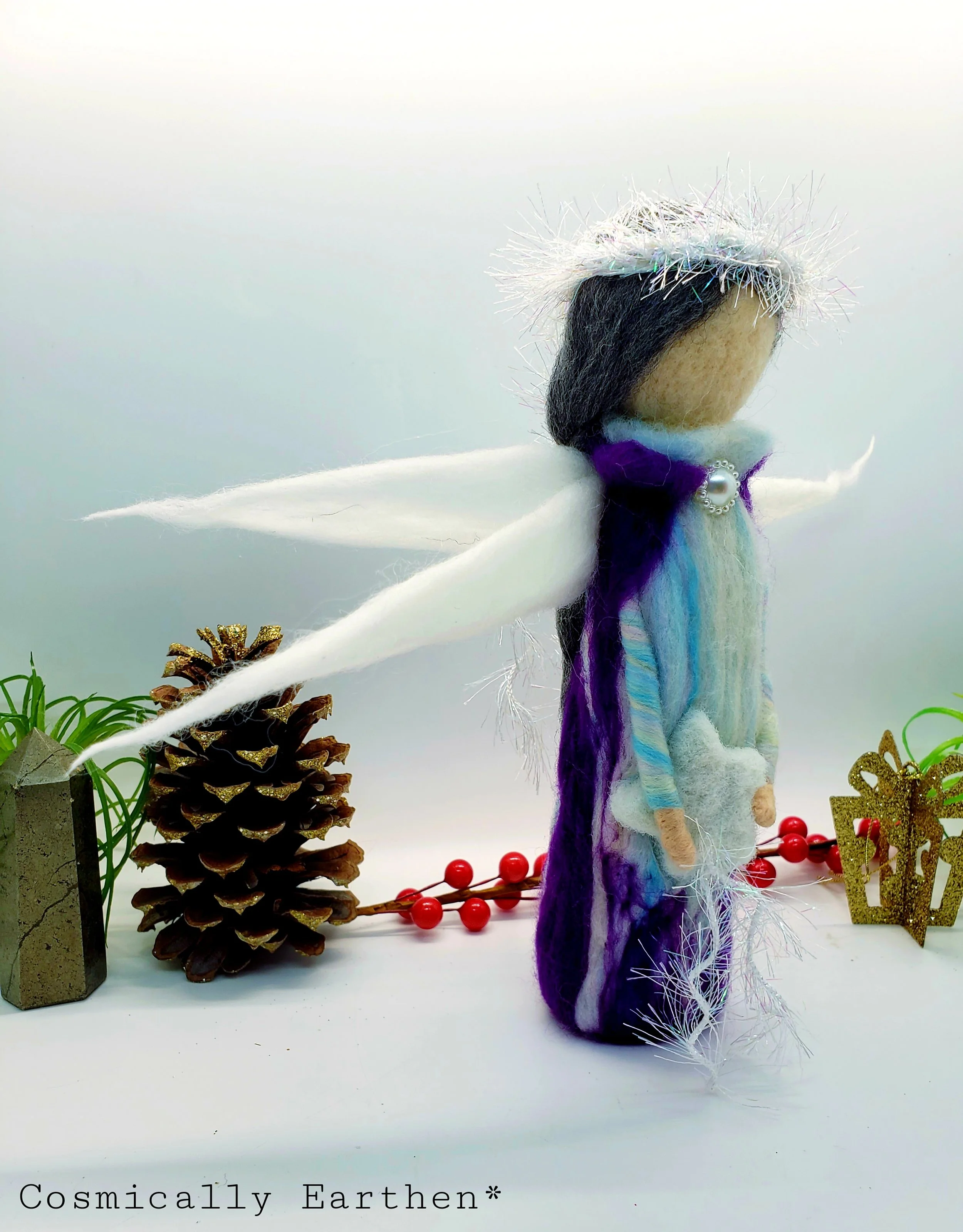 Handcrafted needle-felted angel doll standing in profile, with soft white wings, a shimmering halo, and flowing blue and purple wool garments, styled with natural elements including a pinecone, greenery, and festive accents.