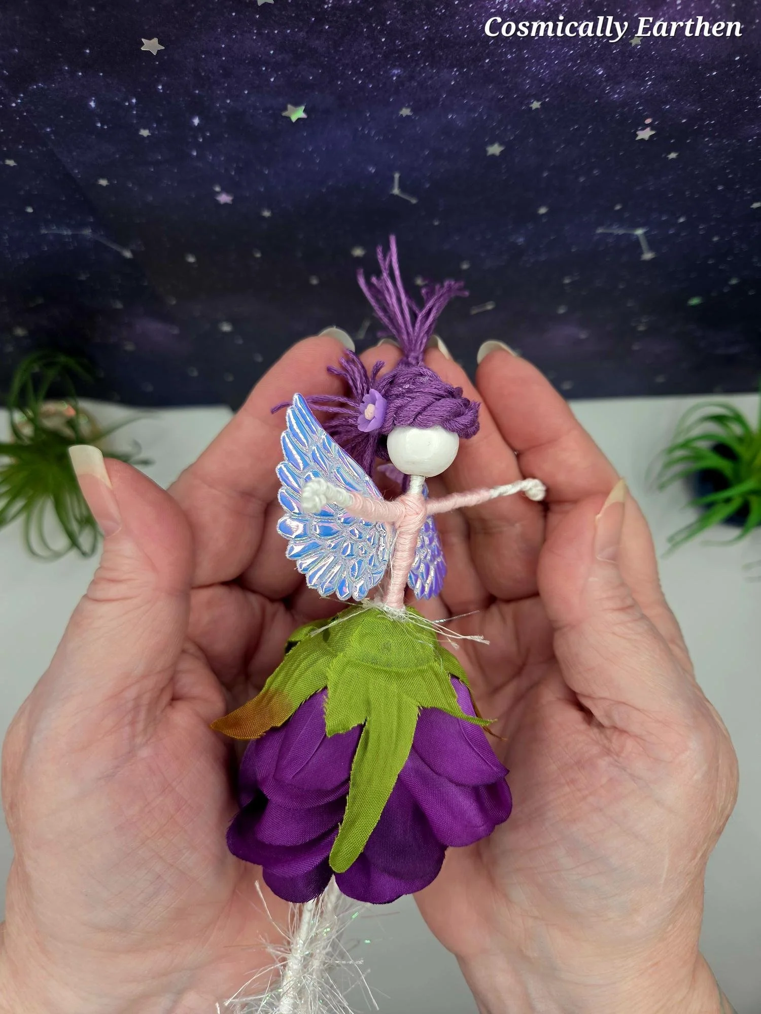 A handcrafted fairy doll with purple hair, iridescent blue wings, and purple fabric clothing, being held by hands in front of a starry night sky background with the text 'Cosmically Earthen' at the top.