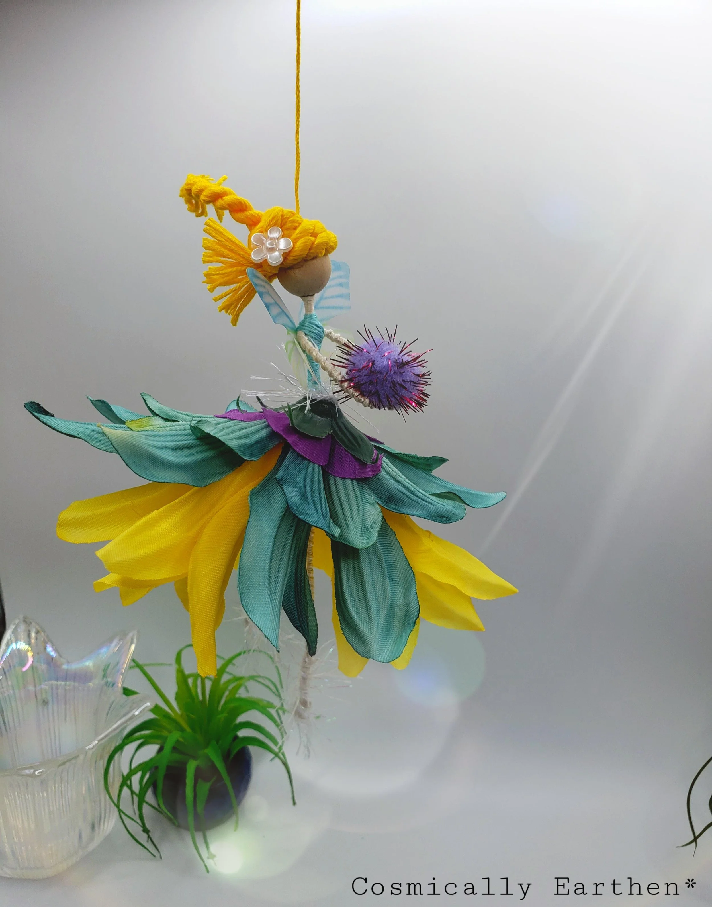 Decorative fairy ornament hanging above a vase with green leaves, made of fabric and yarn in bright colors.