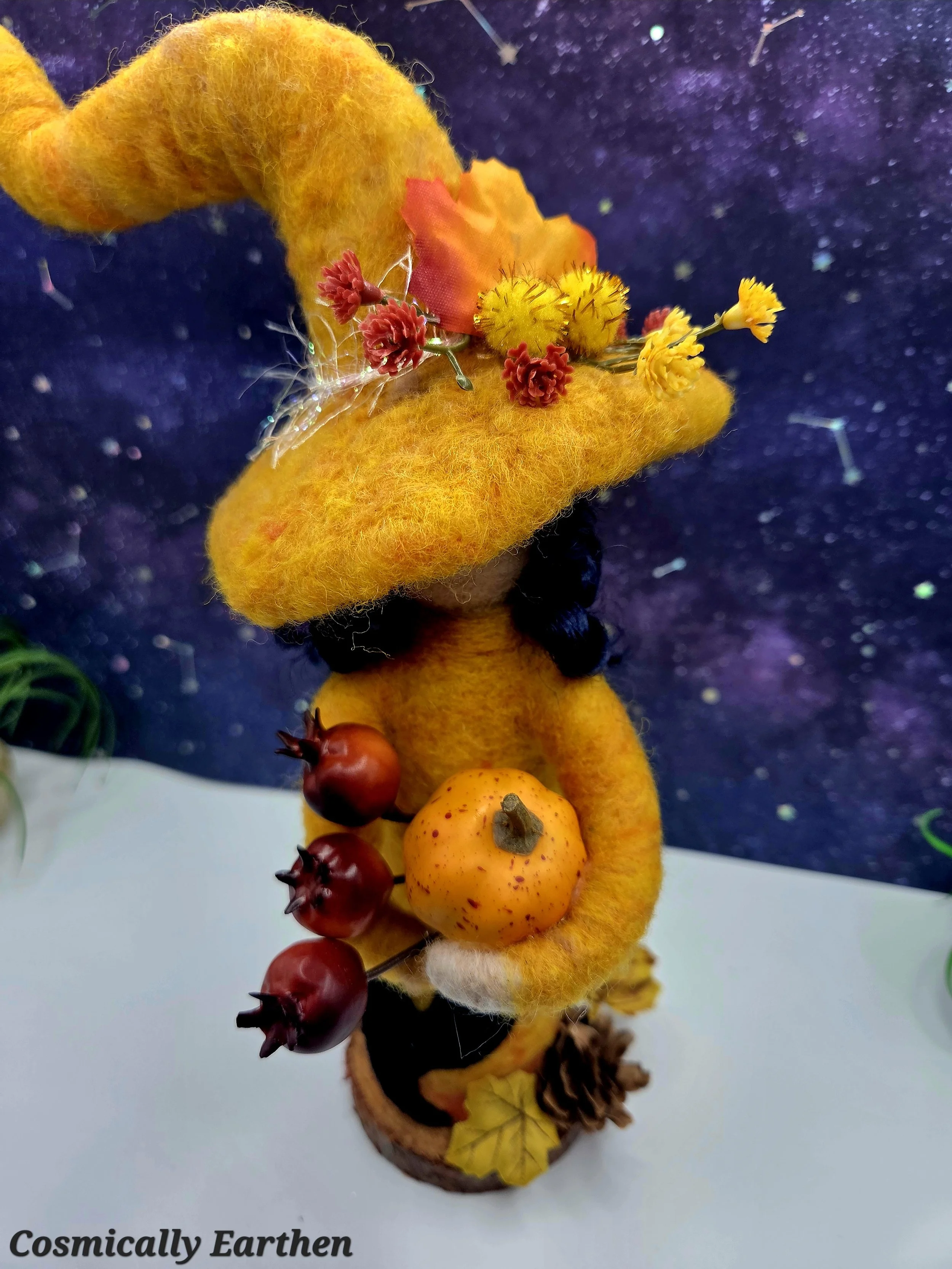 Close-up of needle felted autumn witch doll Marigolde holding harvest fruits including a pumpkin and pomegranates, showing detailed wool textures and handcrafted fiber art features