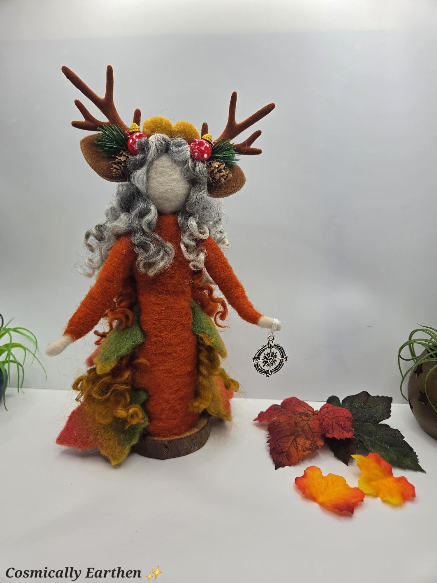 Felt art figure of a reindeer with antlers decorated with Christmas ornaments, pinecones, and greenery, dressed in a costume with orange and green felt. The scene includes autumn leaves and a potted plant, with the caption 'Cosmically Earthen' and sparkles.