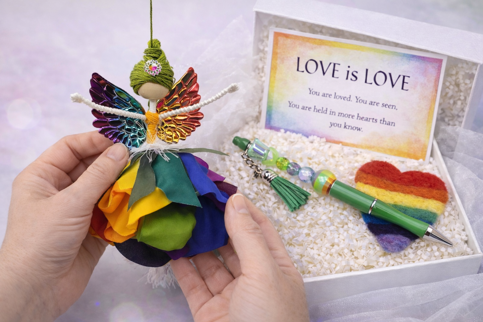 A person holding a rainbow-colored butterfly ornament with fabric flowers, placed in a white gift box filled with shredded paper. The box contains a colorful beaded keychain, a green pen with a rainbow-colored heart-shaped felt decoration, and a card with the message 'LOVE is LOVE' in rainbow colors.