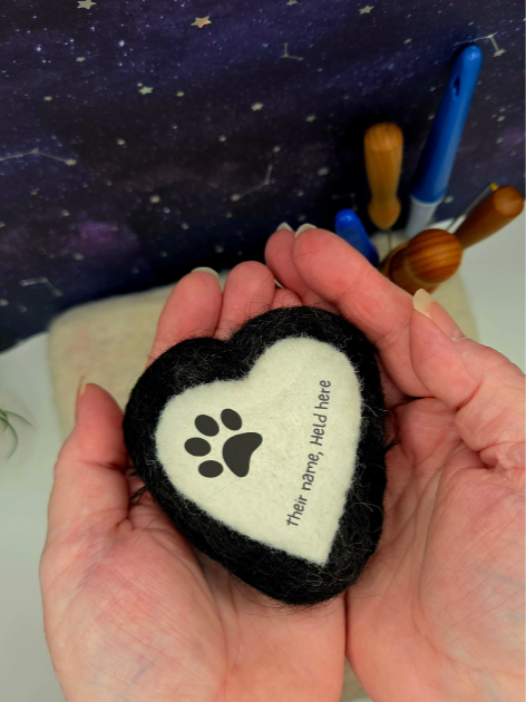 A person holding a heart-shaped felt object with a paw print and customizable text on it, in front of a starry sky background with pens and pencils.