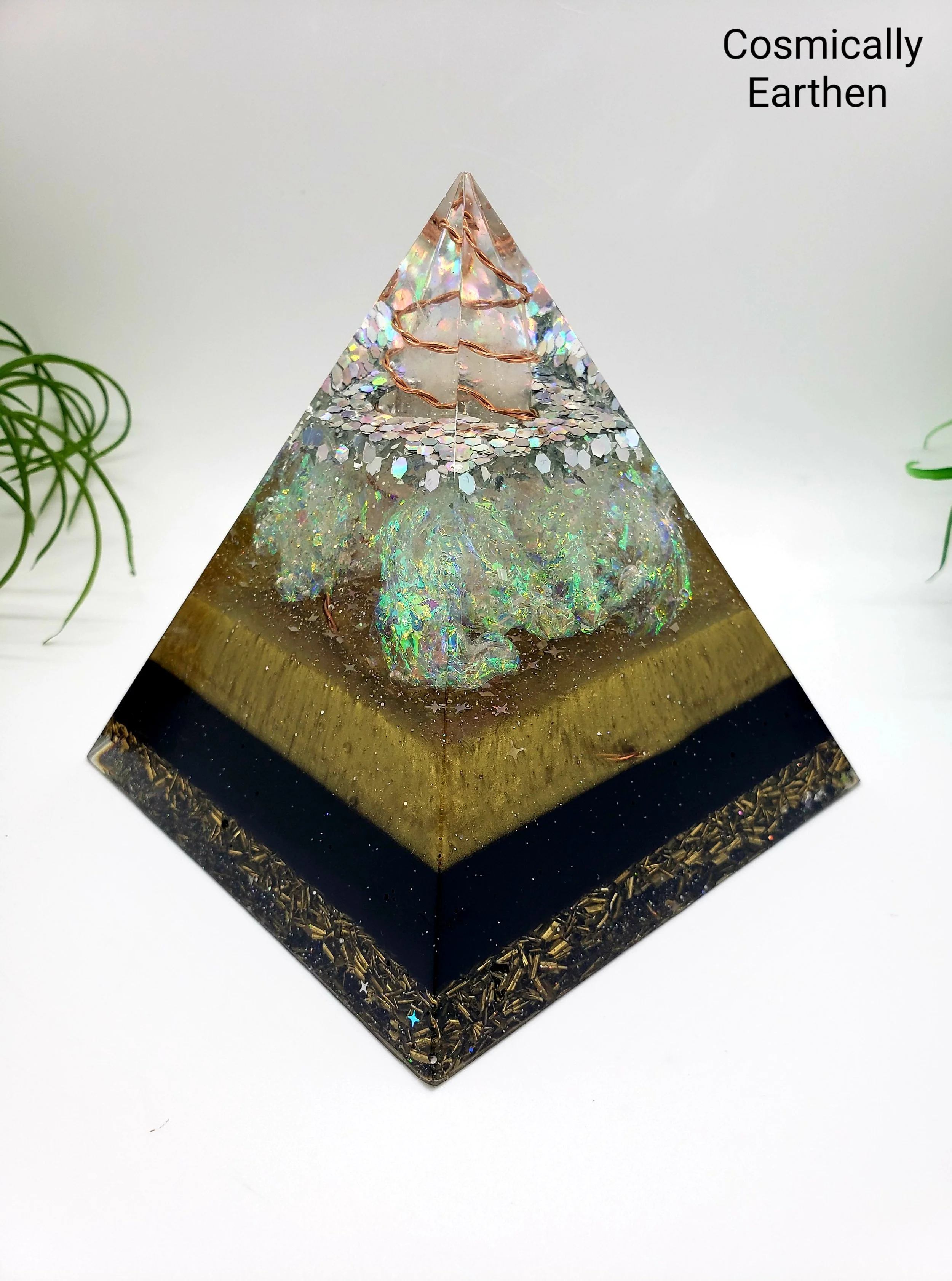A decorative pyramid-shaped glass sculpture with iridescent colors, decorated with rose gold wire, rests on a black base with gold accents, set against a white background with green plants on the sides.