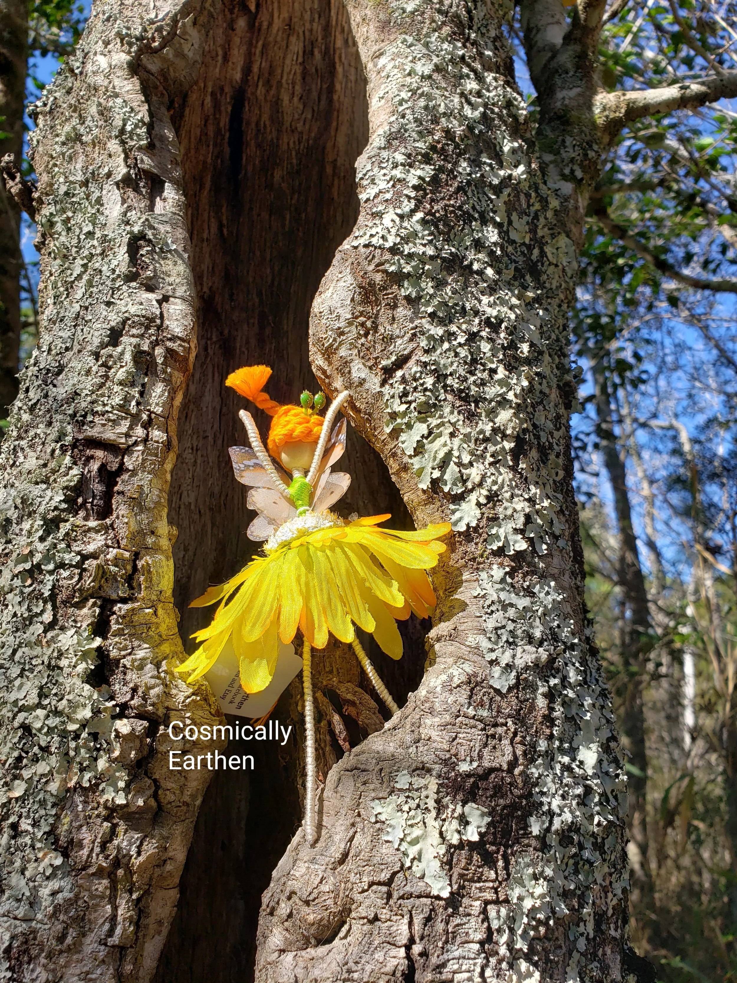 A decorative fairy figurine with yellow, orange, and white features, perched inside a tree hollow with gray bark and lichen, against a background of blue sky and trees.