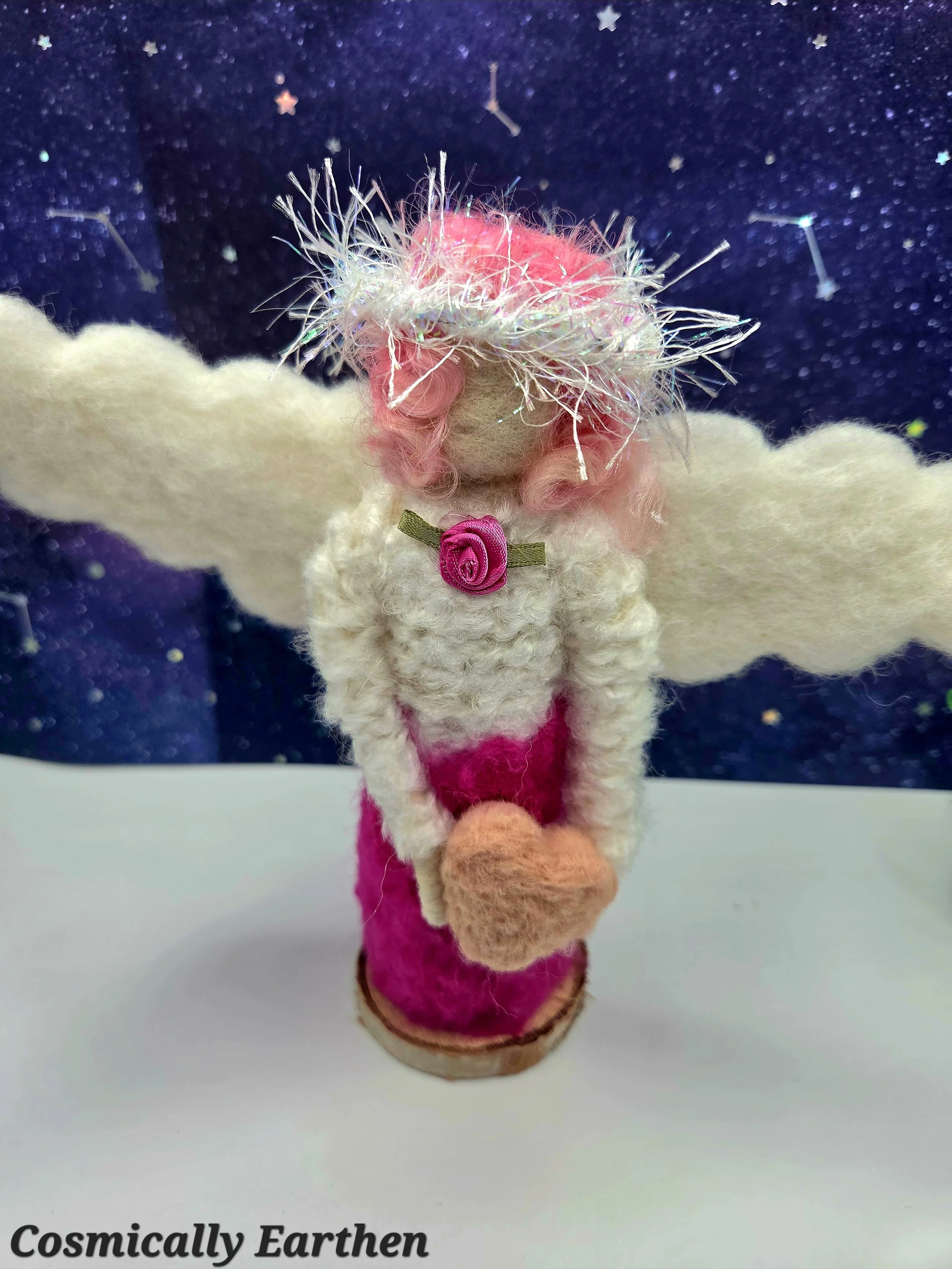 Close-up of handmade needle felted angel holding a small felt heart, featuring soft pink curls, cream wings, and textured wool details, set against a celestial background