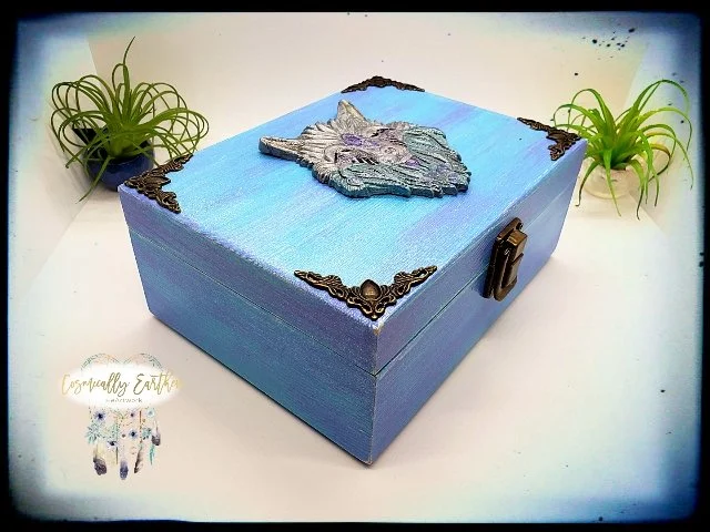A rectangular blue box with black corner accents and a latch on the front, decorated with a 3D mountain and lake scene on the top, and flanked by green plants on either side.