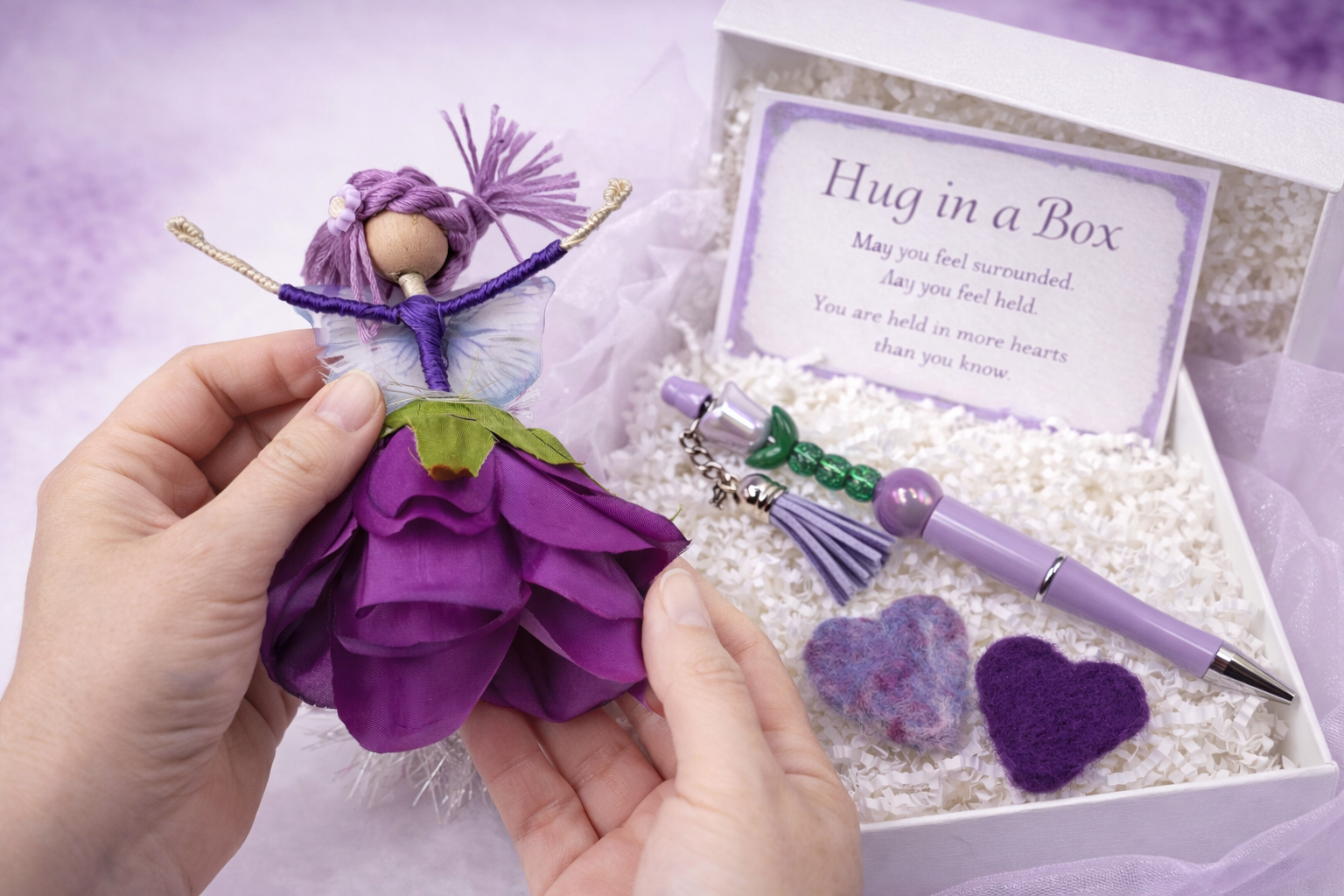 A person holding a small purple fairy doll with purple hair and wings, wearing a purple dress made of fabric. The doll is in front of a white gift box filled with shredded paper, hearts, a lavender pen, and a beaded keychain with a tassel. A card inside the box reads 'Hug in a Box' with a message about feeling surrounded and held in hearts.