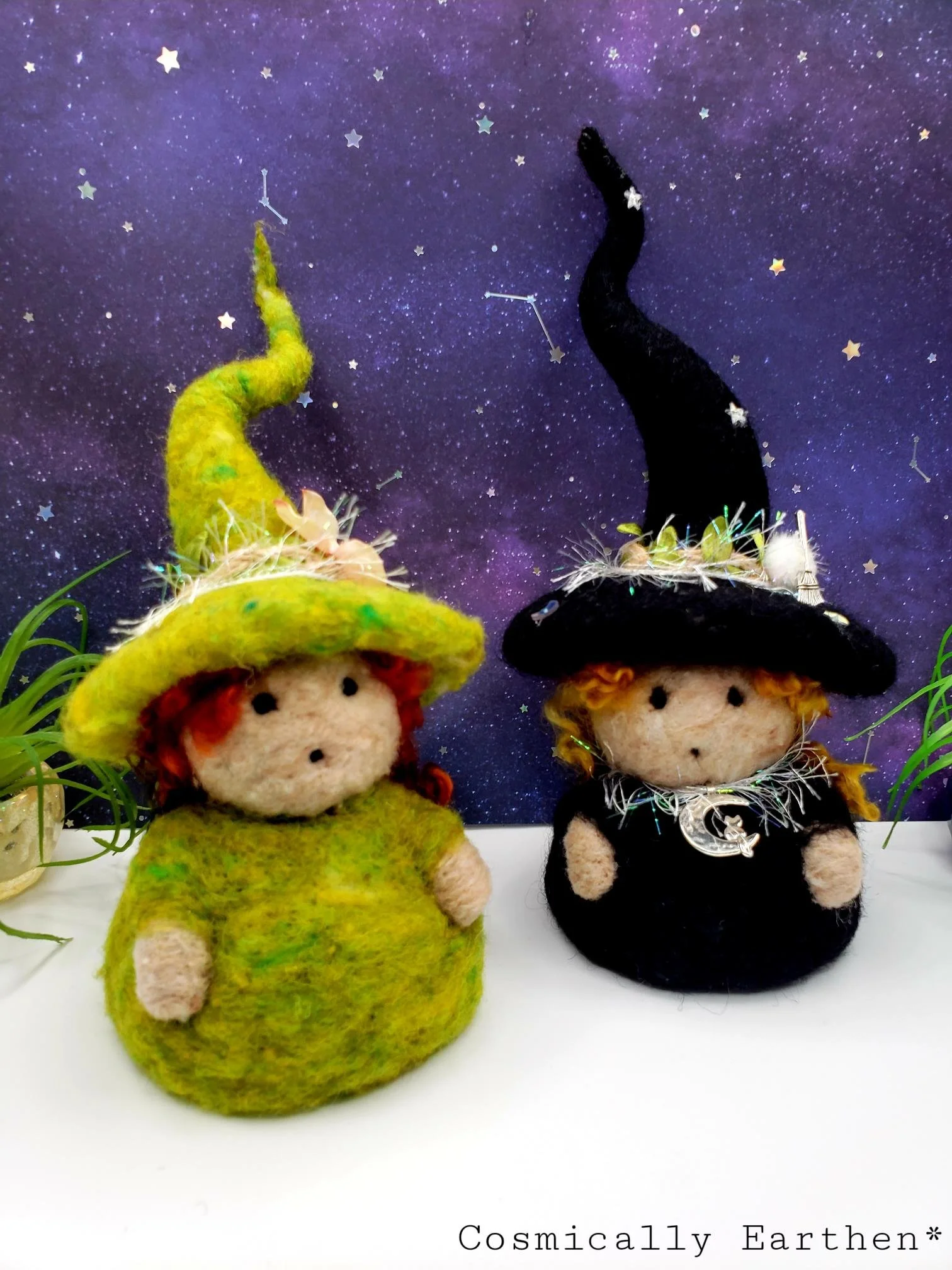Two handcrafted felt dolls dressed as witches with pointed hats, one green and one black, against a cosmic starry background with small decorative stars.