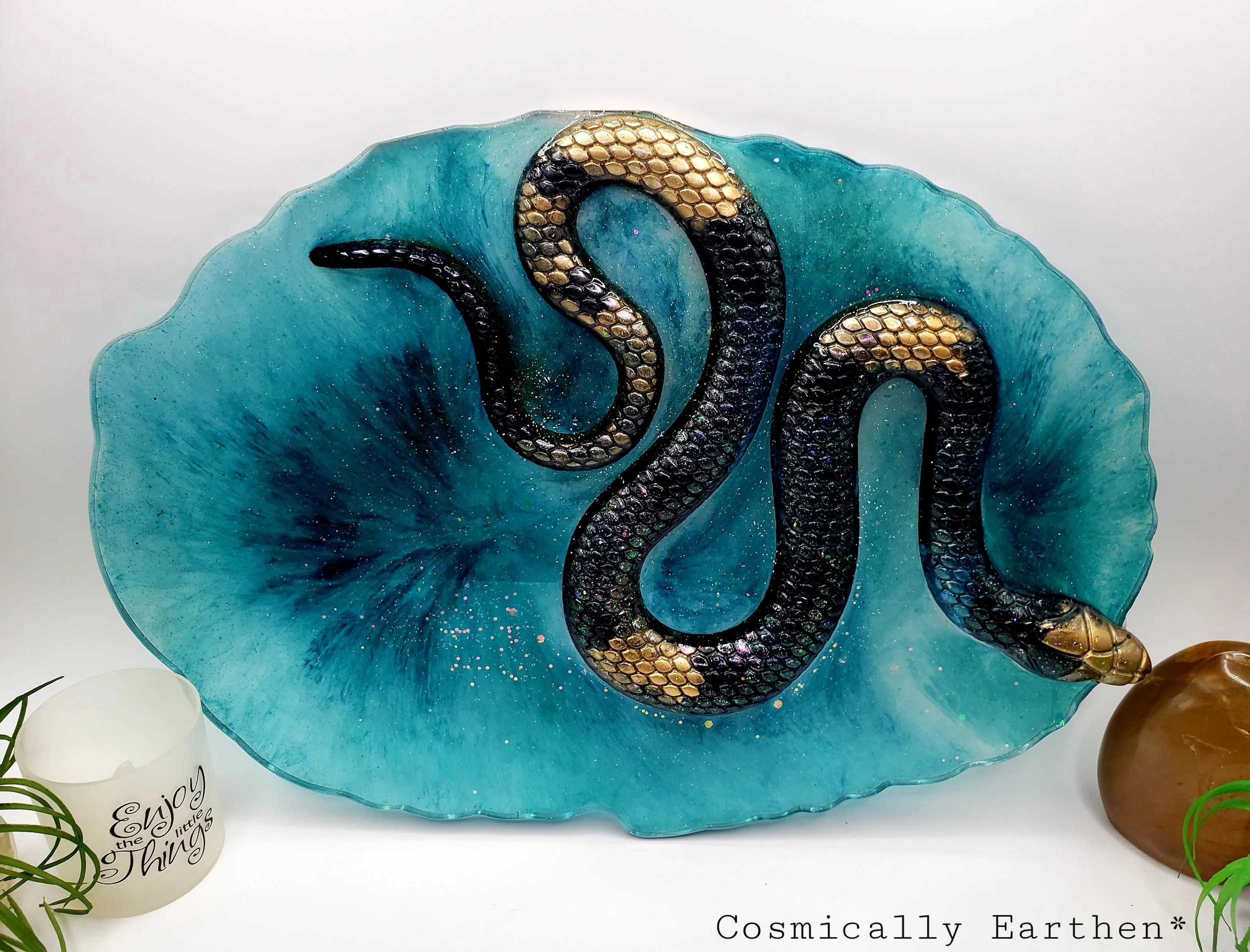 Handcrafted resin geode tray in vibrant blue with a black and gold snake design. Crystals rest on the tray, which is used for displaying sacred items. Surrounded by plants and mystical decor.