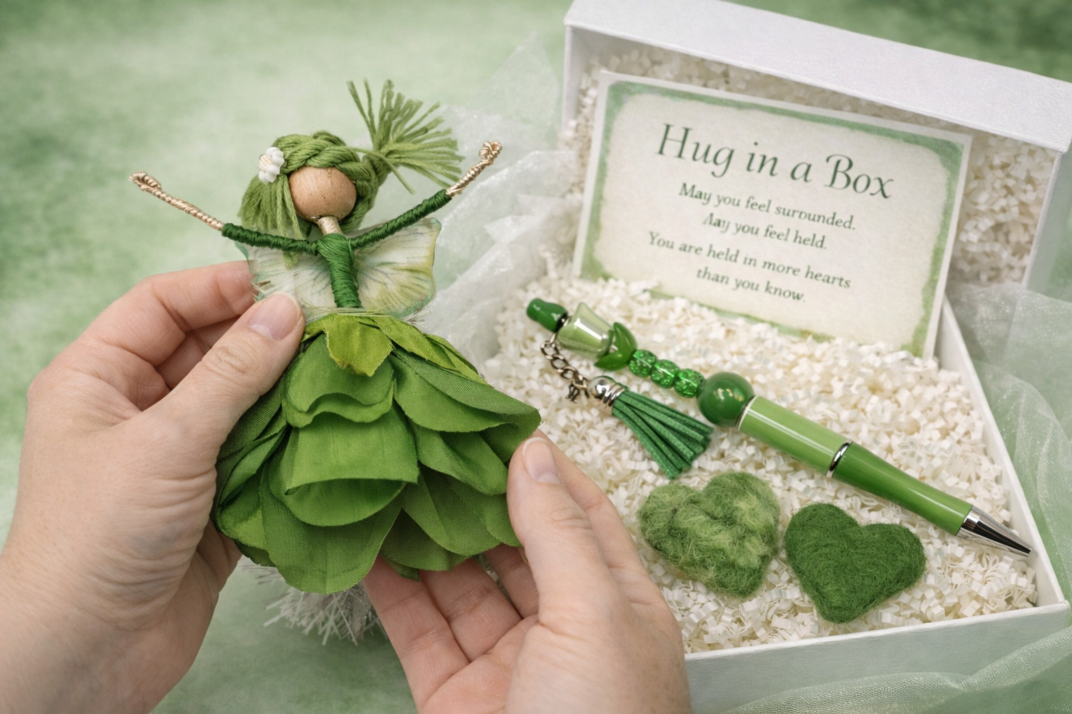 A person holding a small fairy doll with green fabric dress and green hair in front of a decorative box containing green heart-shaped felt ornaments, a green pen, a necklace, and a card titled 'Hug in a Box' with a poem inside.