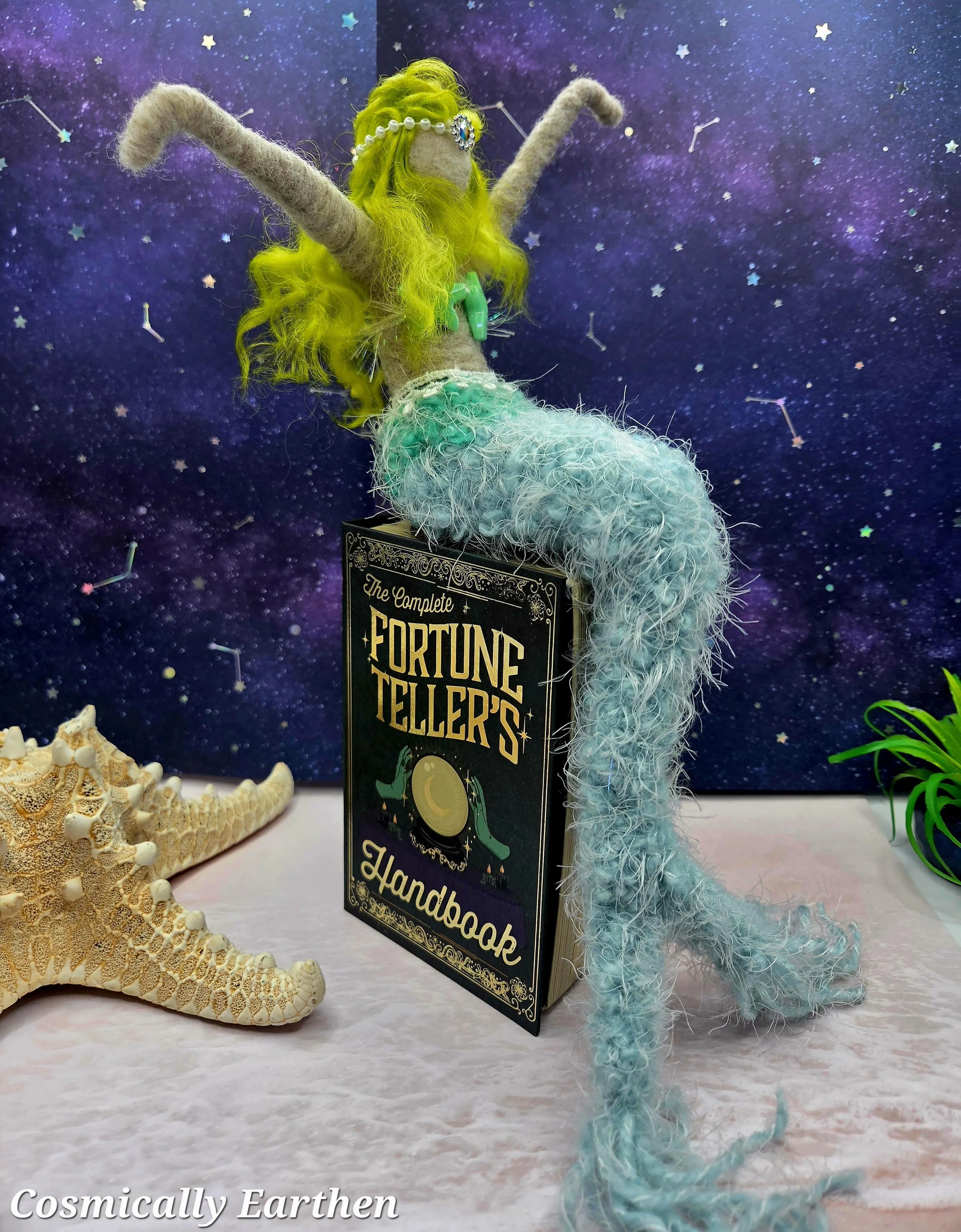 A whimsical mermaid doll with long yellow hair, a pearl headband, and a turquoise gemstone necklace, sitting on a book titled 'The Complete Fortune Teller's Handbook.' The background features a starry, outer space scene, and there is a starfish on the surface beside the doll.
