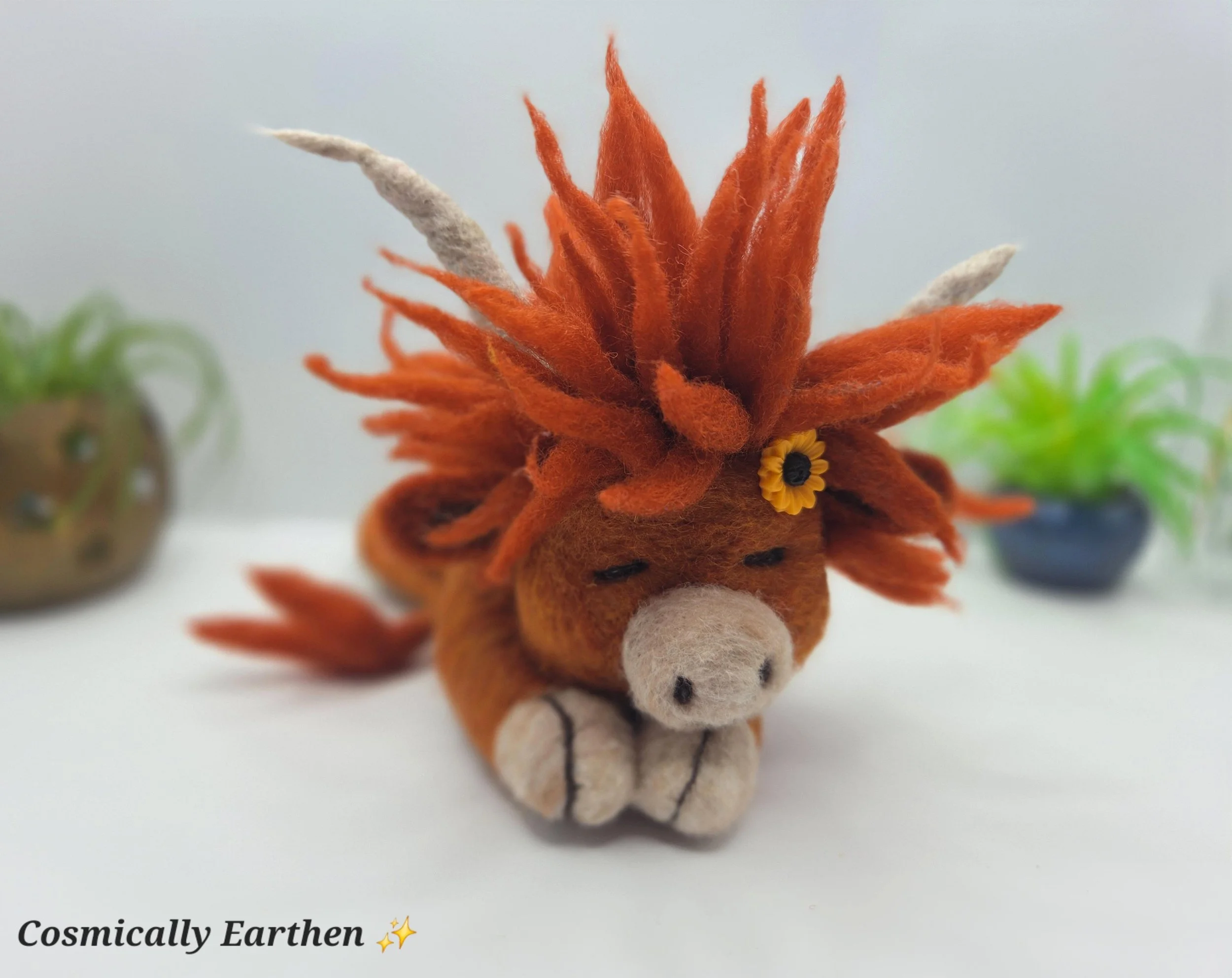 Felted animal figure resembling a fox with orange, white, and brown felted wool, with green and beige felted plants in the background.