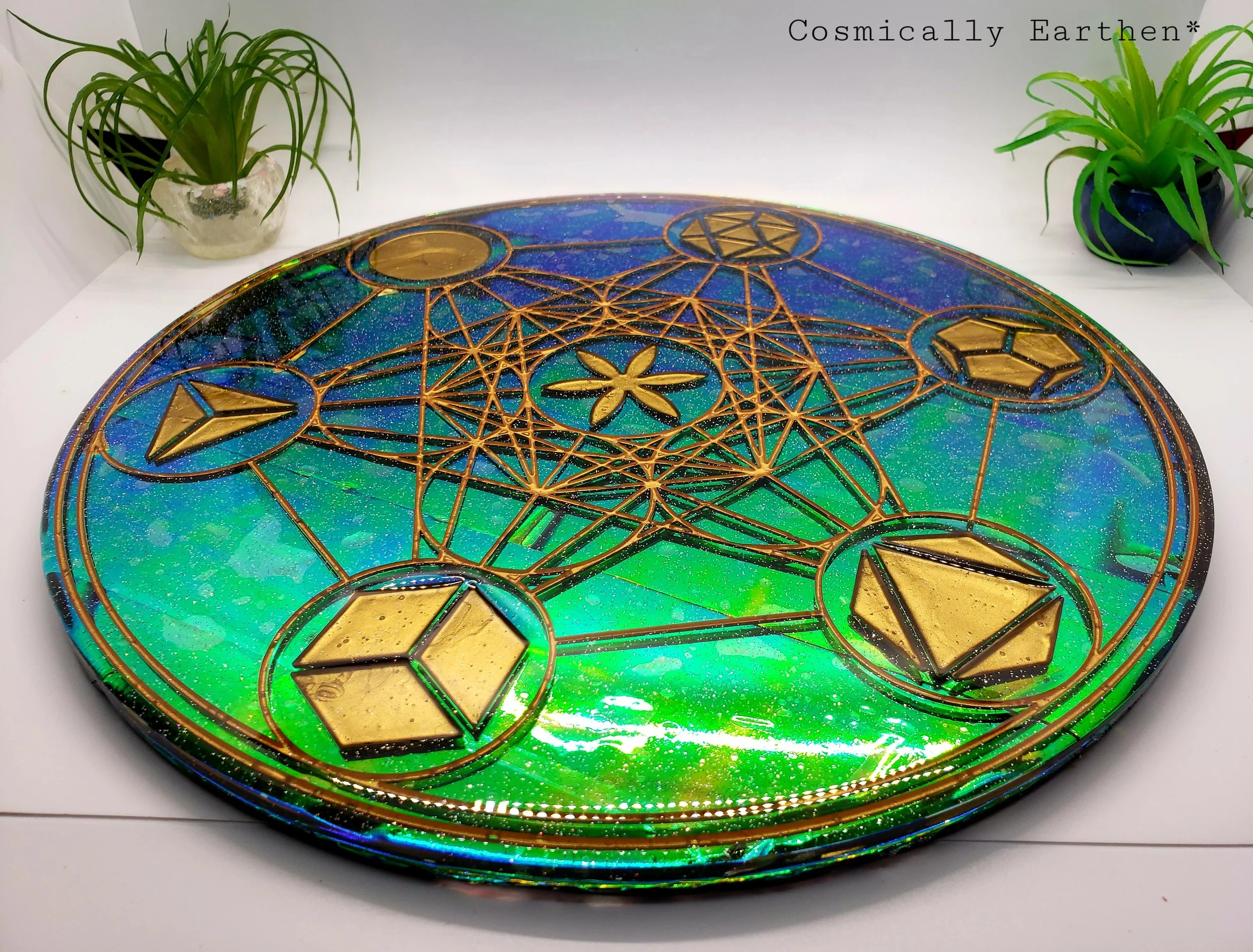A round, colorful glass object with intricate gold geometric patterns and a star design in the center, placed on a white surface with two potted plants in the background.