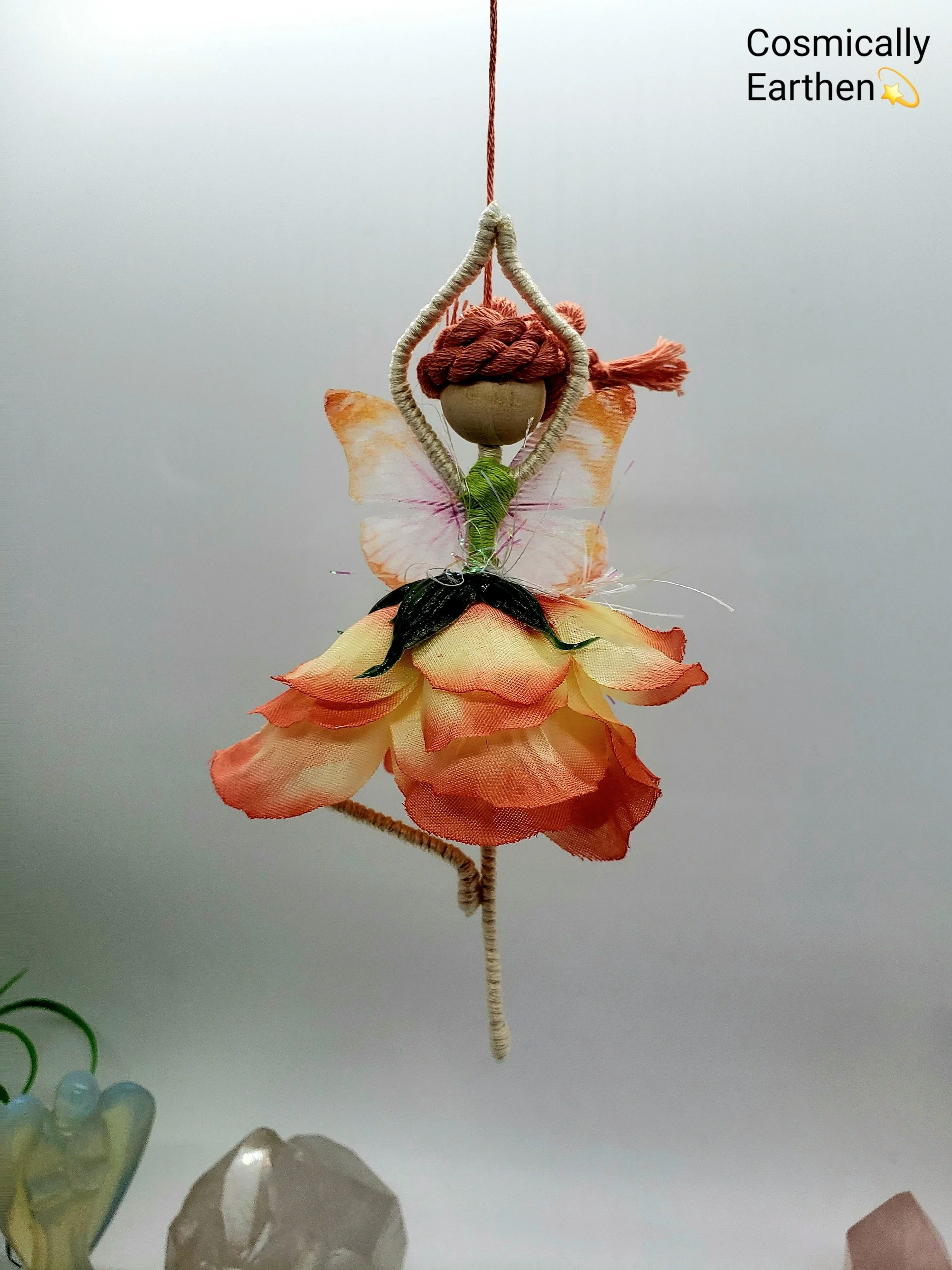 A decorative hanging ornament resembling a fairy or angel, with butterfly wings, a head made from a round object, and a body with fabric flower petals, hanging from a string against a plain background.