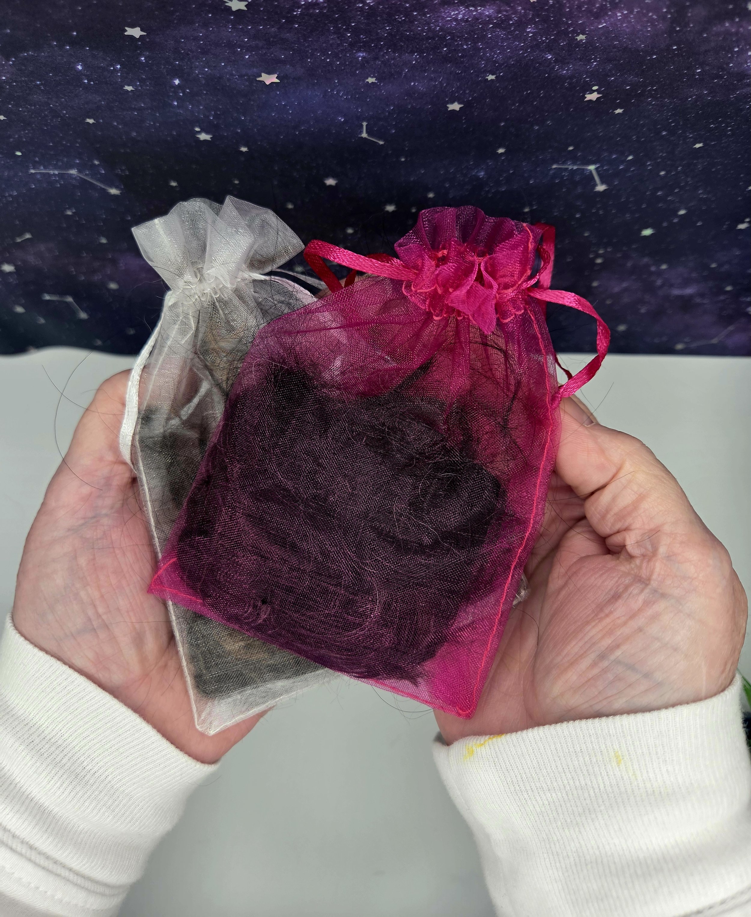 Two small organza gift bags, one silver and one pink, are held in a person's hands. The bags appear to contain small items, possibly jewelry or trinkets. The background features a starry night sky with constellations.