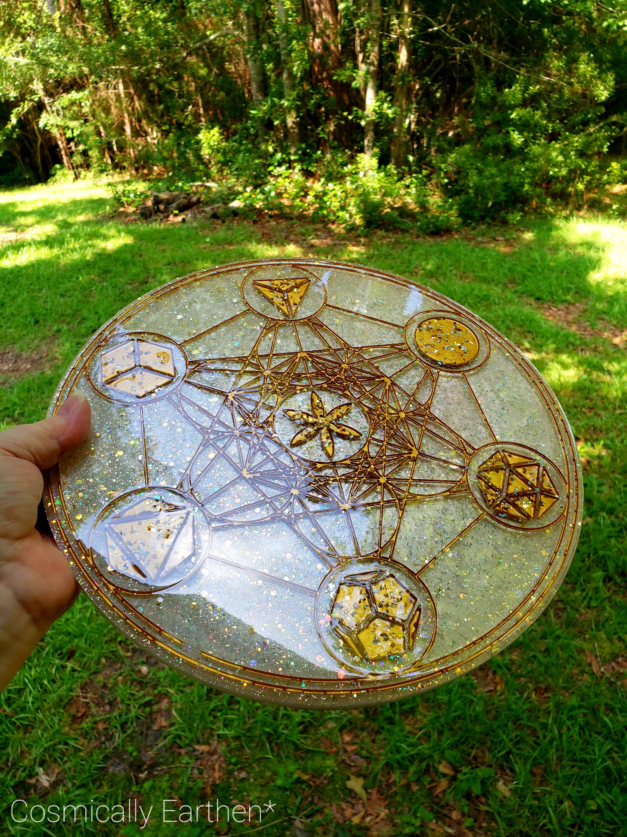 A decorative glass plate with golden geometric and celestial patterns being held outdoors over grass and trees.