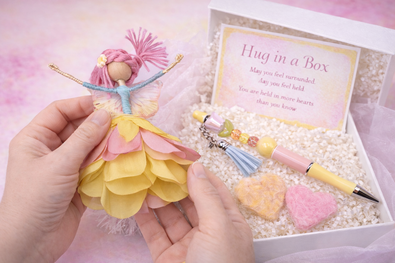 Person holding a fairy doll dressed in a yellow and pink petal-like dress, with a gift box containing a bracelet, a pink and yellow pen, two felt hearts, and a card that reads "Hug in a Box" with a poem.