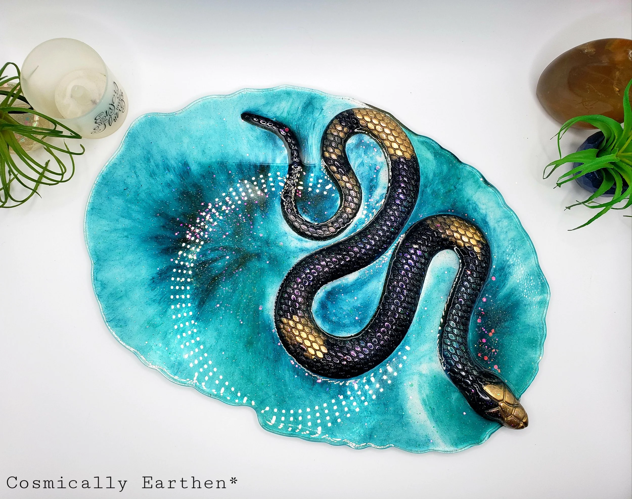 Handcrafted resin geode tray in vibrant blue with a black and gold snake design. Crystals rest on the tray, which is used for displaying sacred items. Surrounded by plants and mystical decor.