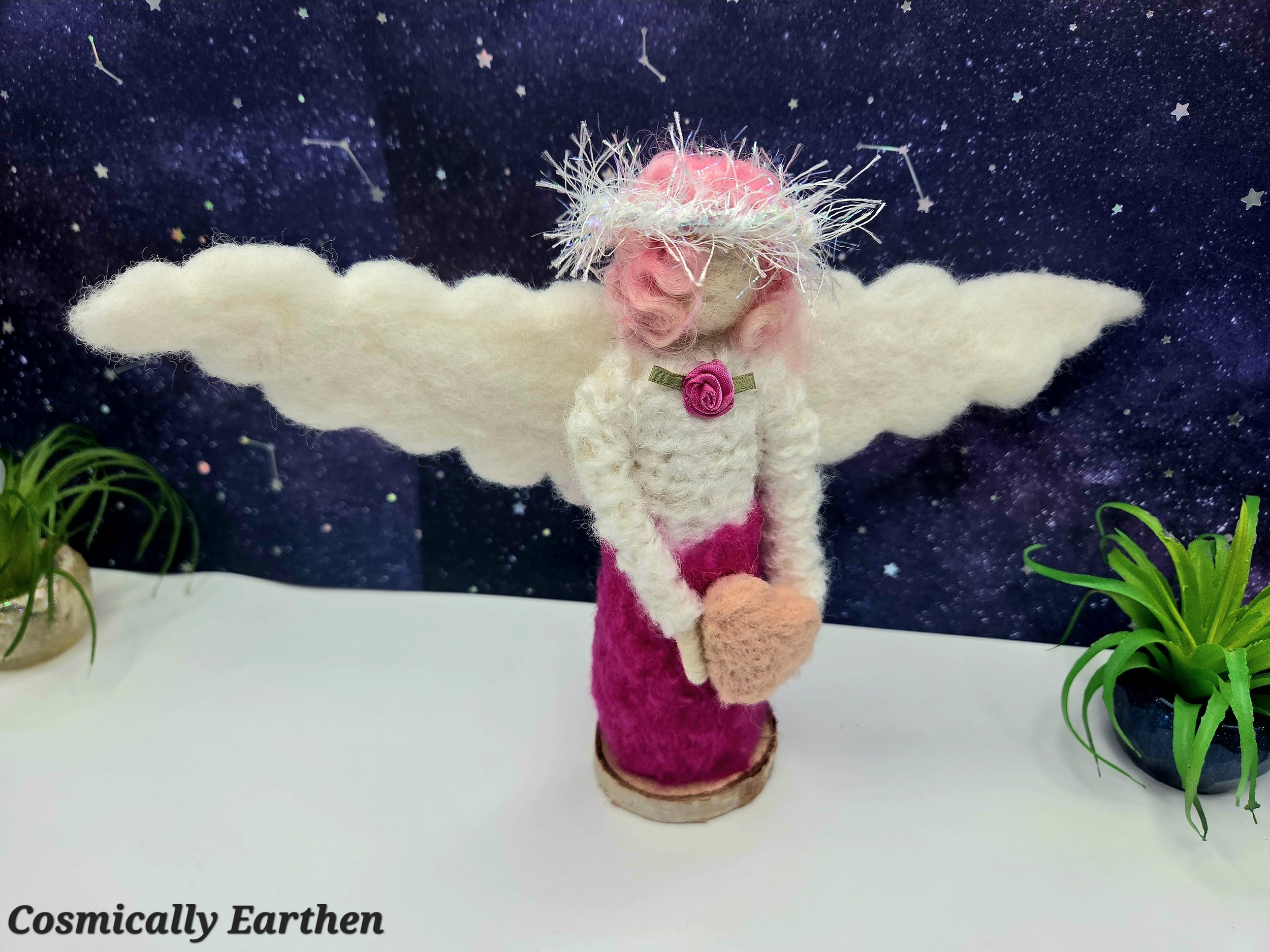 Handcrafted needle felted angel figurine with pink dress, cream wings, and heart in hands, standing on a natural wood base with a starry sky backdrop