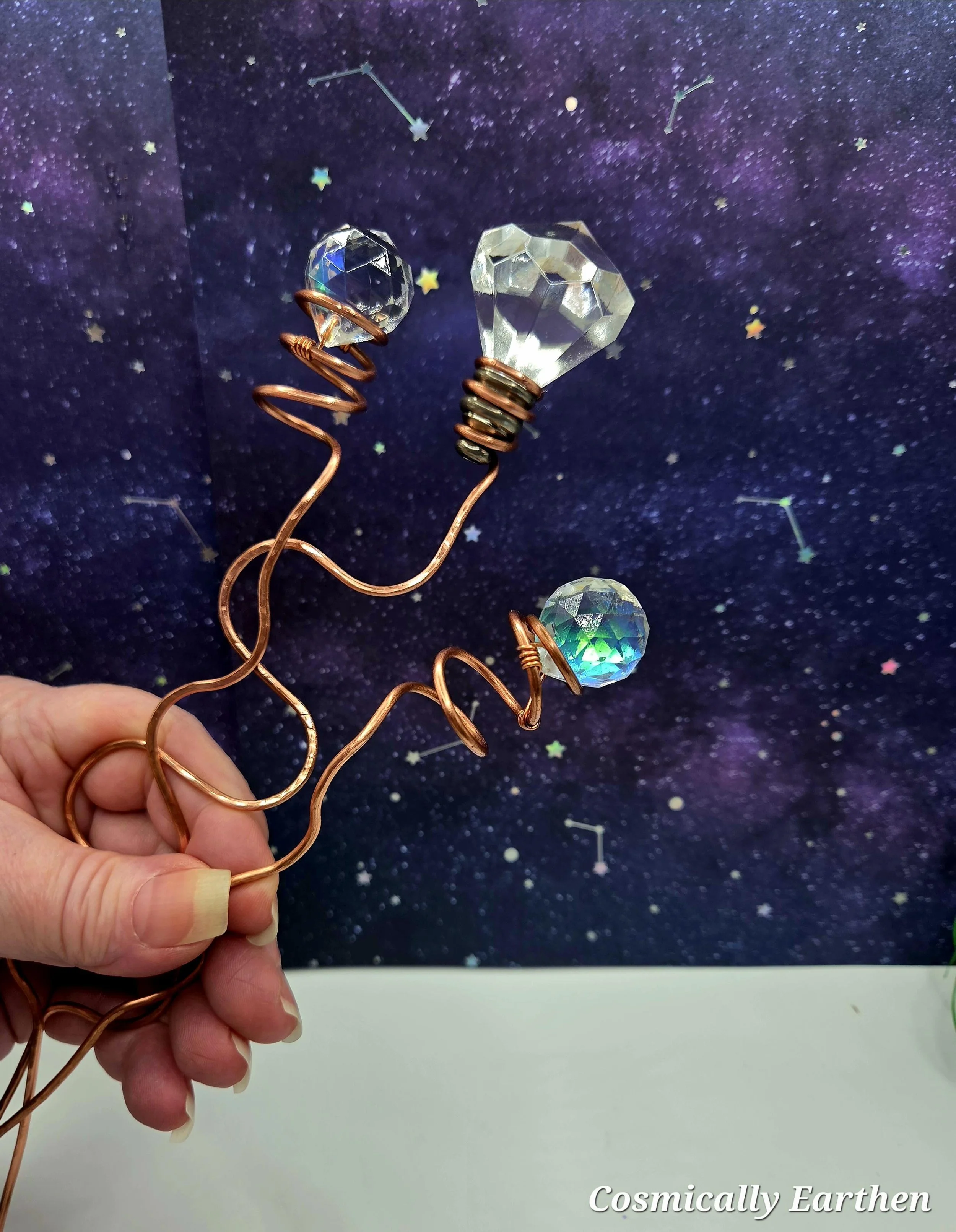 Hand holding two wire jewelry pieces with faceted crystal beads shaped like hearts and circles, set against a cosmic galaxy background with stars and constellations.