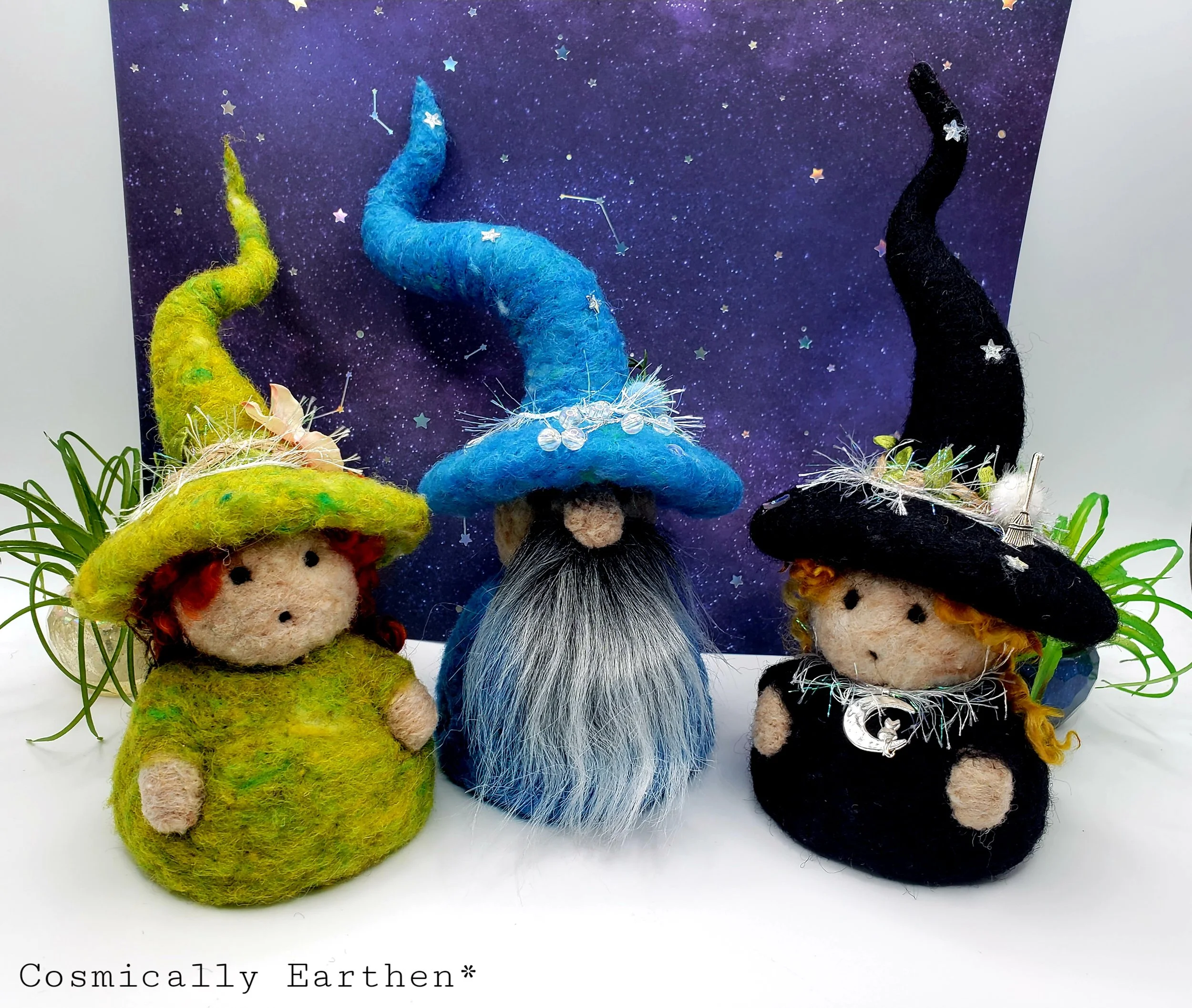 Three felted witch dolls with pointed hats, set against a starry night background. The dolls have different colored hats: yellow, blue, and black, with decorative elements. They are placed with some green plants in the background.