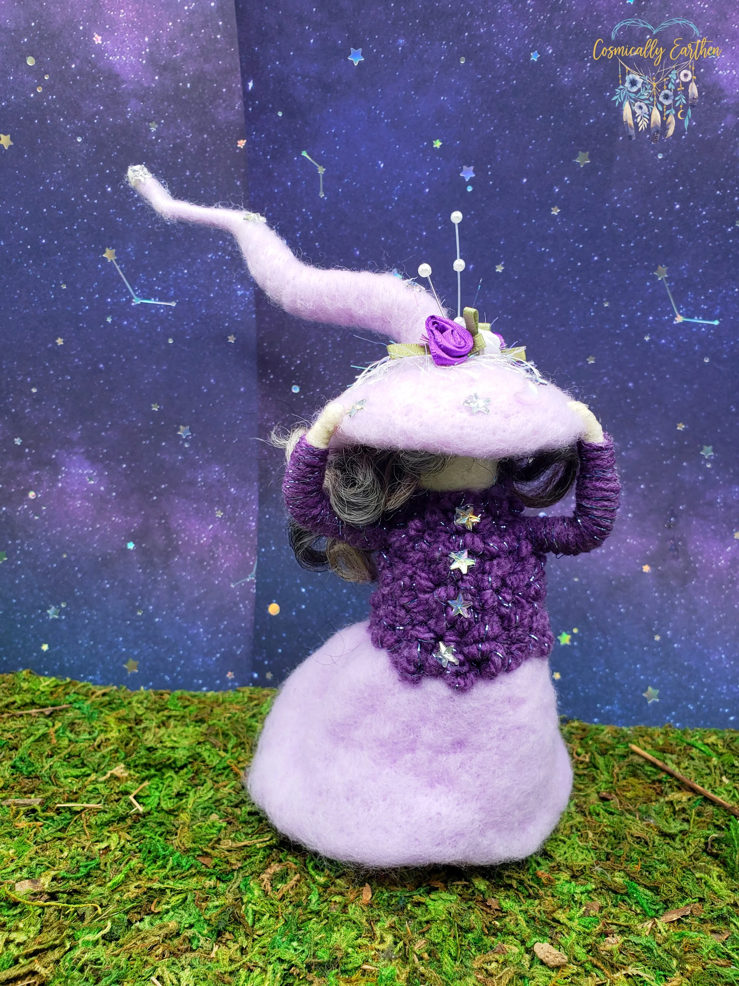 A felted fairy-like creature with a purple and pink color scheme stands on green moss, against a cosmic starry background decorated with constellation stickers and a logo reading 'Cosmically Earthen' in the top right corner. The figure has a tall, pointed pink hat with a purple flower, star decorations, and a purple knitted top with star embellishments.