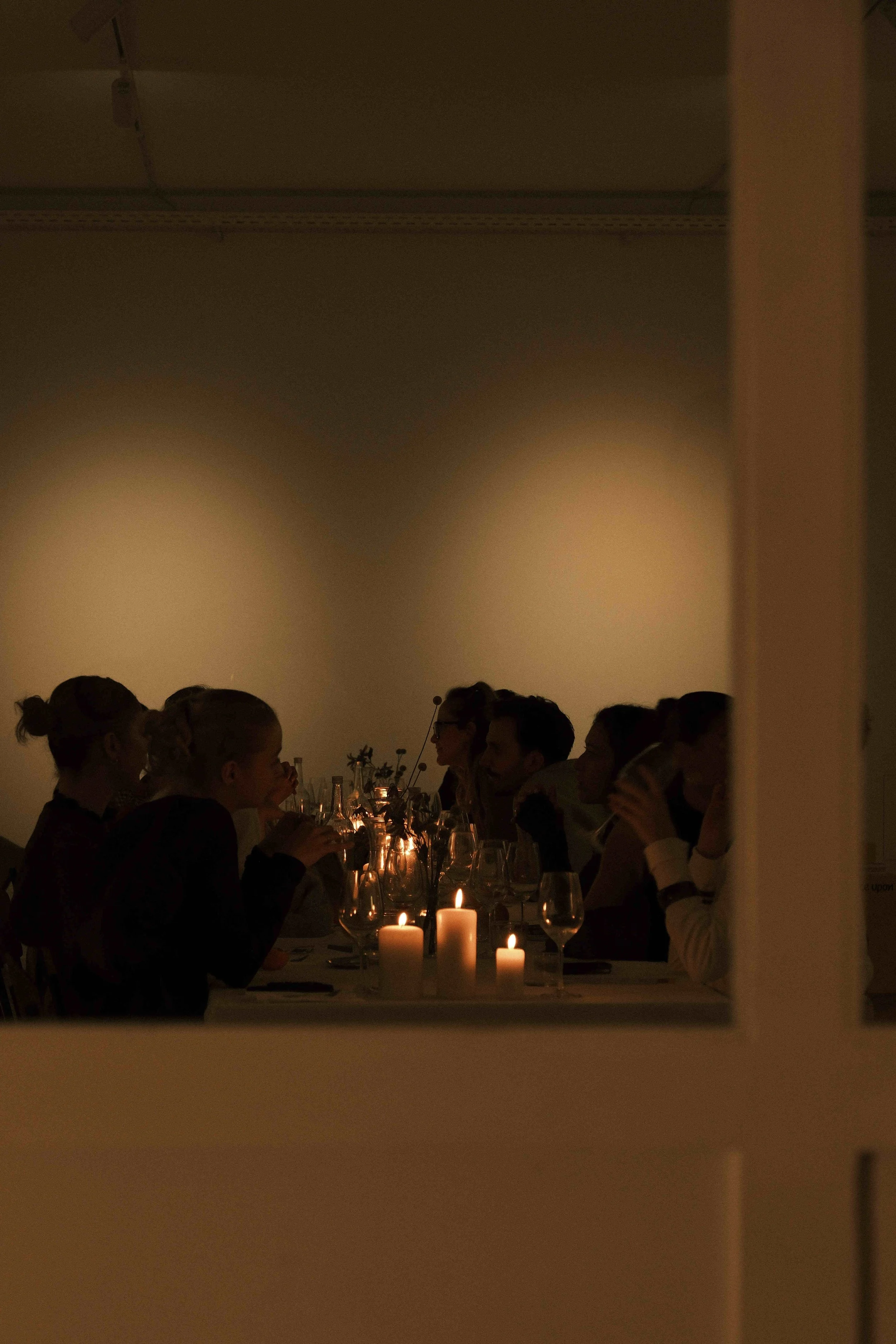 Eine Gruppe von Menschen sitzt an einem Esstisch bei Kerzenlicht, sichtbar durch eine Fensteröffnung, im Dunkeln. Es sieht aus wie eine Abendgesellschaft oder ein Abendessen.