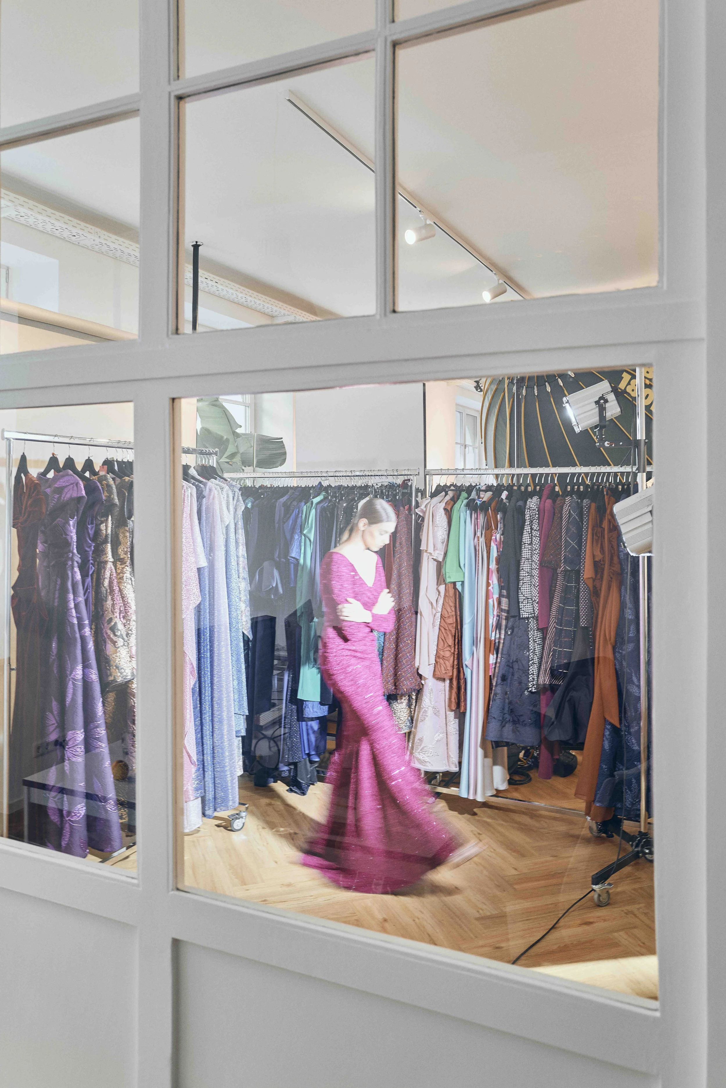 Blick durch eine Glaswand auf eine Modenschau mit einer Model auf dem Laufsteg, umgeben von Kleiderständern mit bunten Kleidern in einem hell erleuchteten Raum.