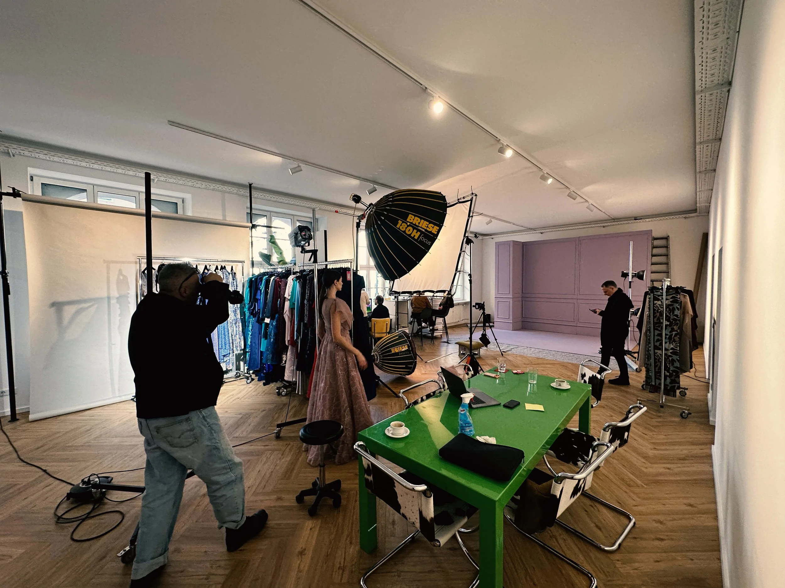Innenaufnahme eines Fotostudios mit Kleidung, Lichttechnik, einem Model in einem Abendkleid, einem Fotografen, einem Stylisten, einem Besprechungstisch und verschiedenen Requisiten.