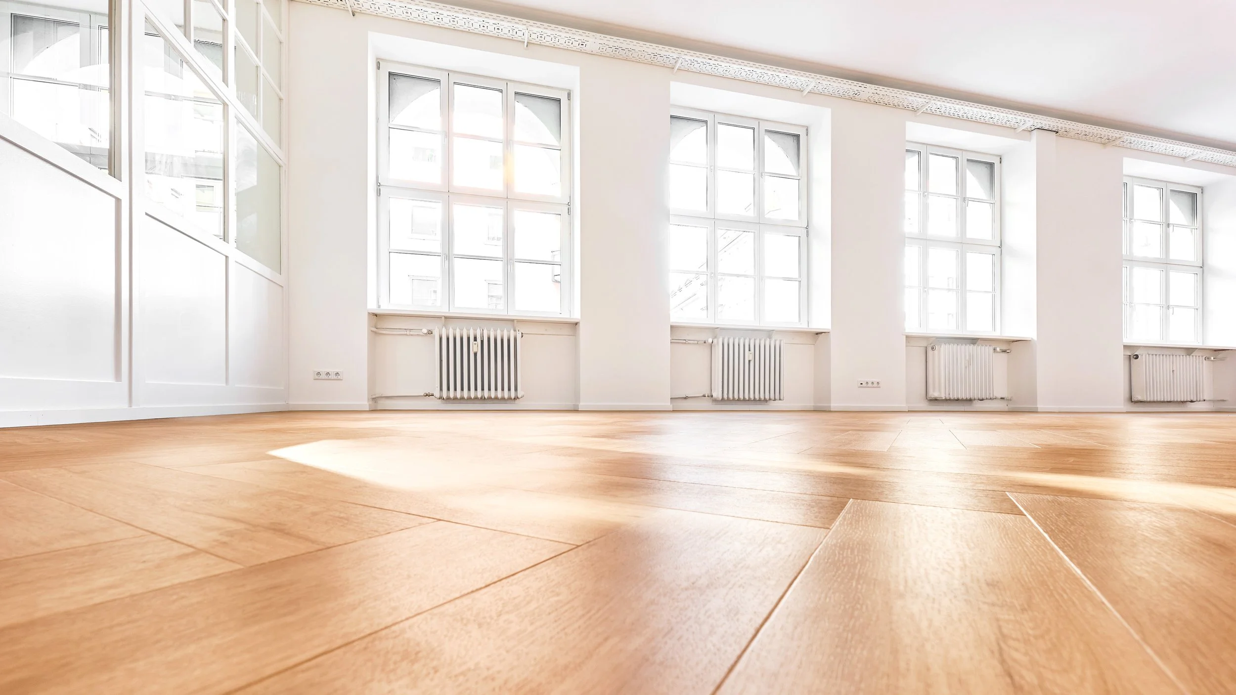 Ein heller, leerer Raum mit großen Fenstern, weißen Wänden und Heizkörpern darunter, Holzfußboden und viel Tageslicht.