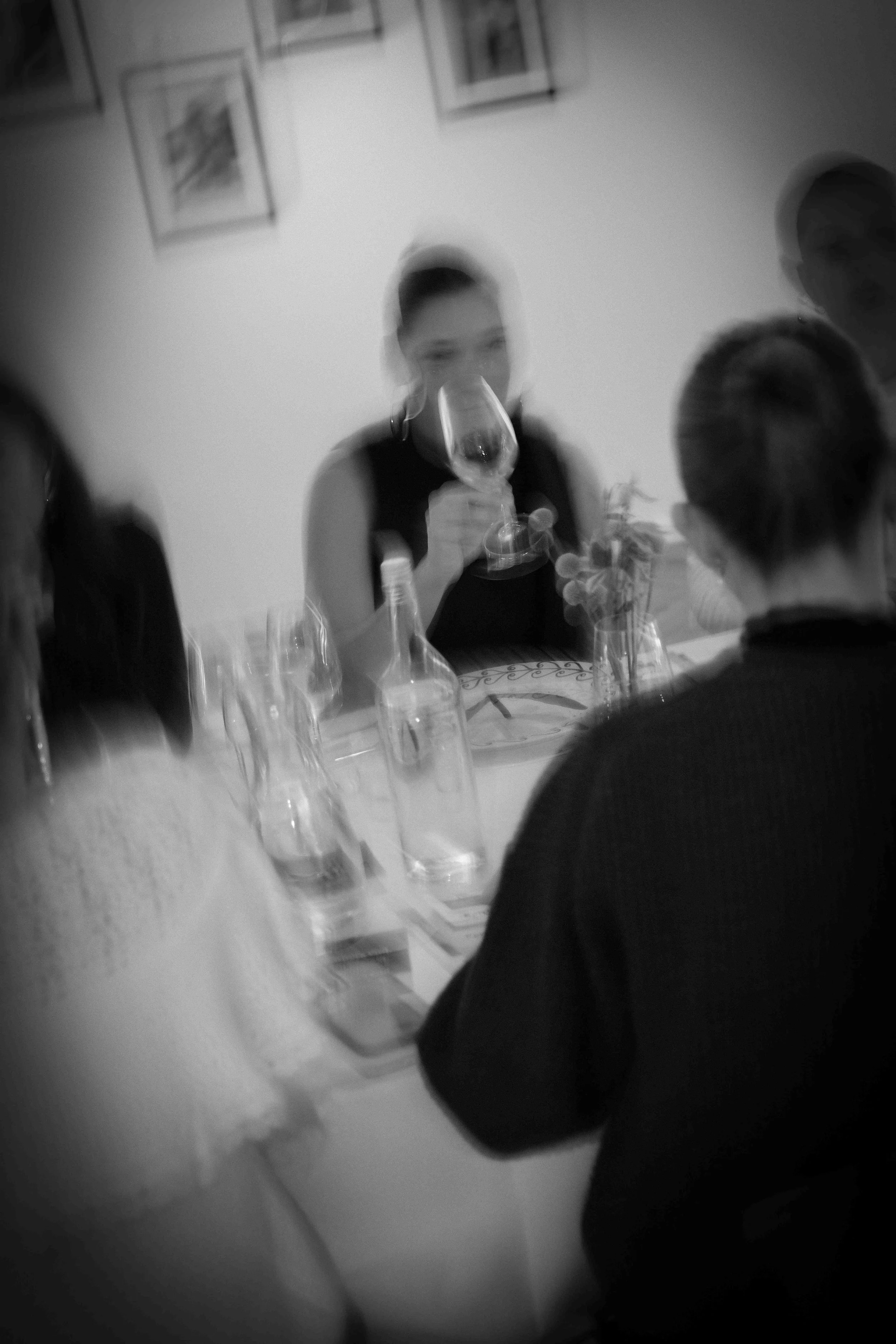Menschen sitzen um einen Tisch bei einem Abendessen, eine Person trinkt Wein, im Hintergrund sind Bilder an der Wand. Die Aufnahme ist unscharf.